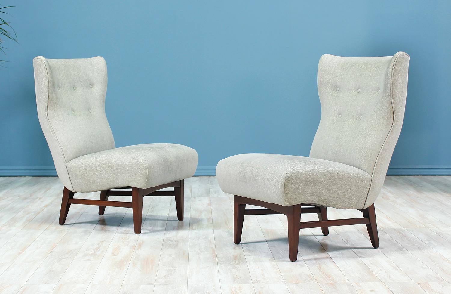 Very comfortable pair of wingback lounge chairs manufactured in the United States, circa 1950s. This lovely pair of lounge chairs features a sturdy walnut wood base and a newly upholstered seat in a beautiful grey hue with tufted details on the