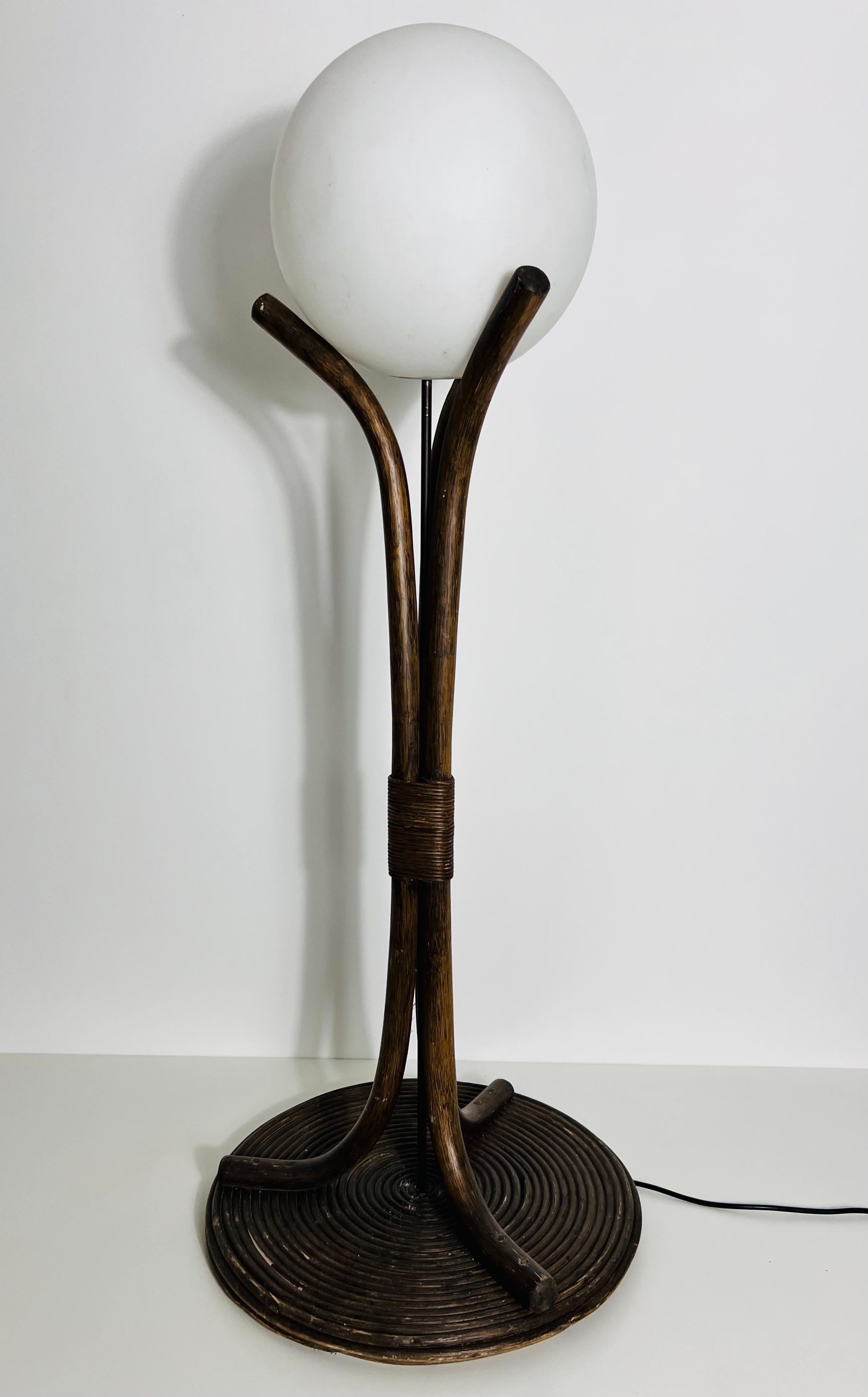 Ce lampadaire, fabriqué en bois de bambou foncé, présente un design où le bois s'ouvre vers le haut. Au sommet, un globe rond en verre opale est maintenu entre les trois bras du support en bois, donnant l'impression que la structure en bambou berce