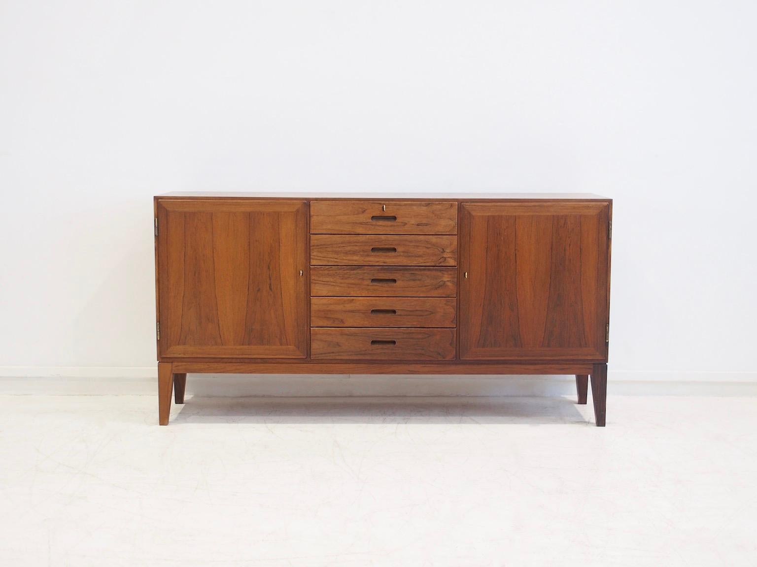 Scandinavian Modern sideboard with slanted legs designed by Kaj Winding. Five drawers with a lock on the top one, keys included. Two doors behind which are shelves. Back of wood veneer, labelled Danish furniture makers control.