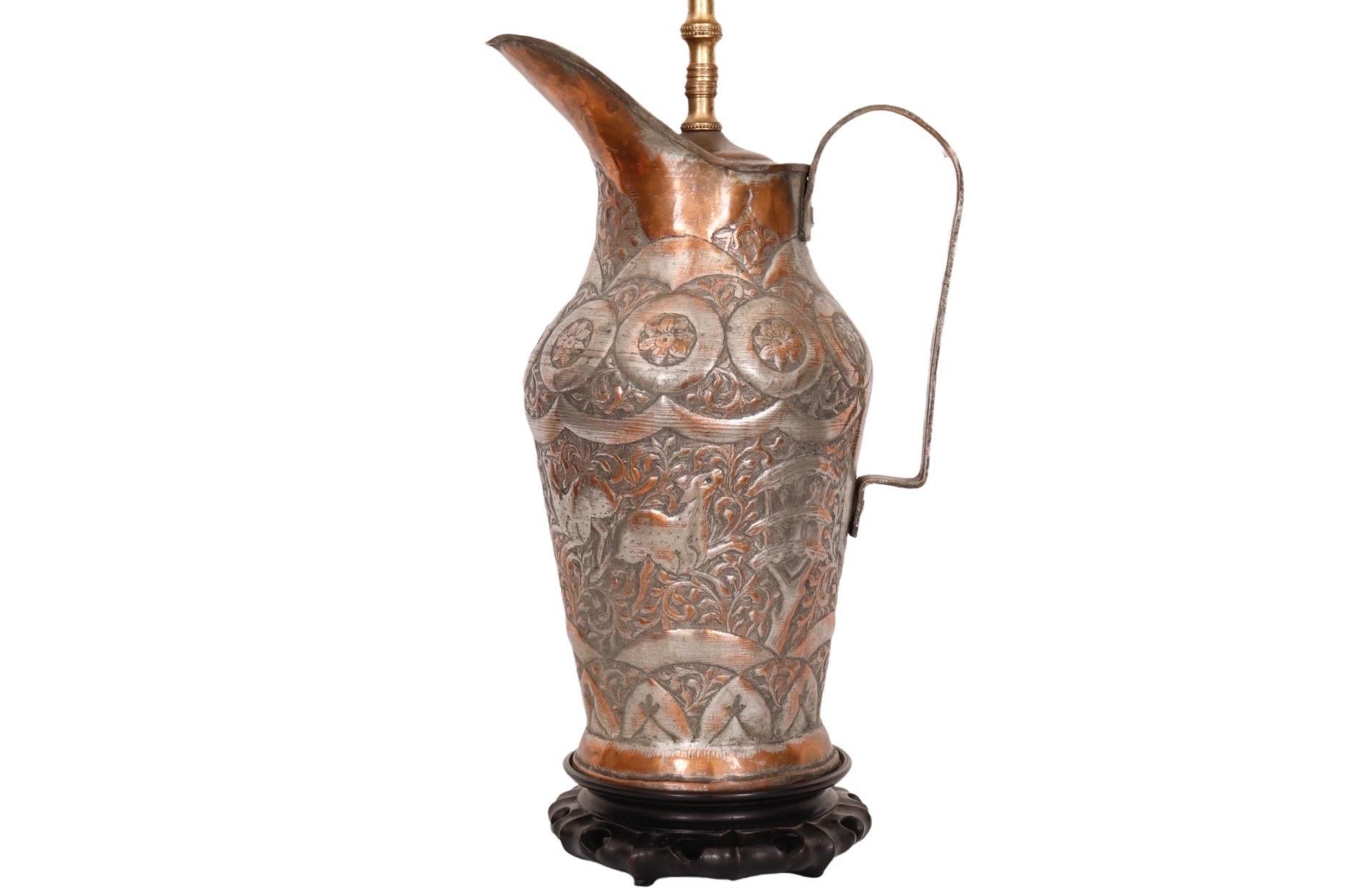 A 19th century Middle Eastern silver over copper and brass water pitcher, converted into a table lamp. The pitcher has a riveted handle and is etched with deer amid vine foliage on the main body, a flower pattern around the neck and a symmetrical