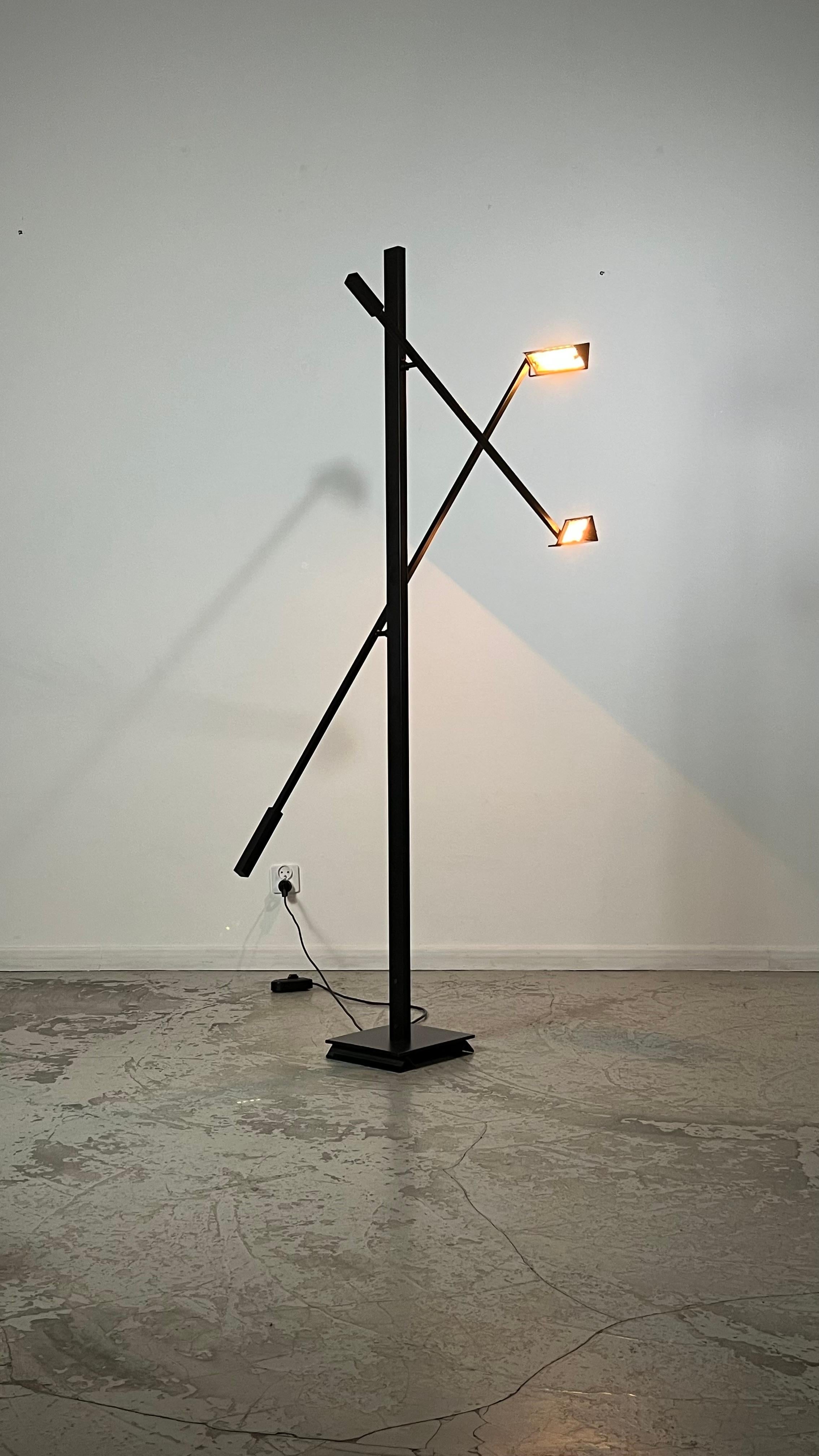 The lamp is constructed out of a black lacquered metal frame rectangle with two adjustable shades.
The pount where the three rods meet creates a set of remivable lines.
Both shades are fitted with a hologen lamp and can be switched on and dimned