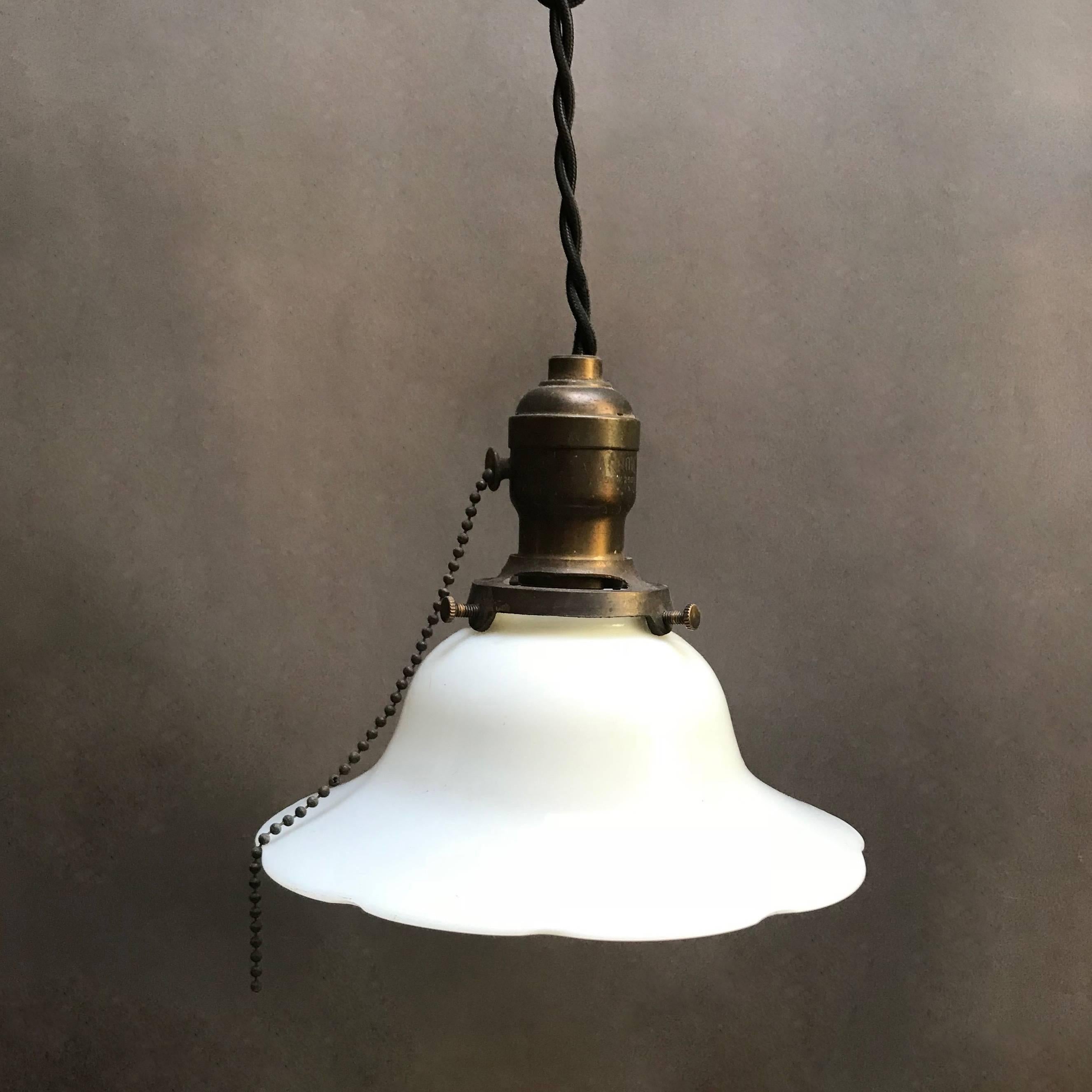 Industrial pendant light features a milk glass, bell shape shade with scalloped edge with brass pull chain fitter is newly wired with 48 inches of braided black cloth cord to accept a 150 watt bulb.