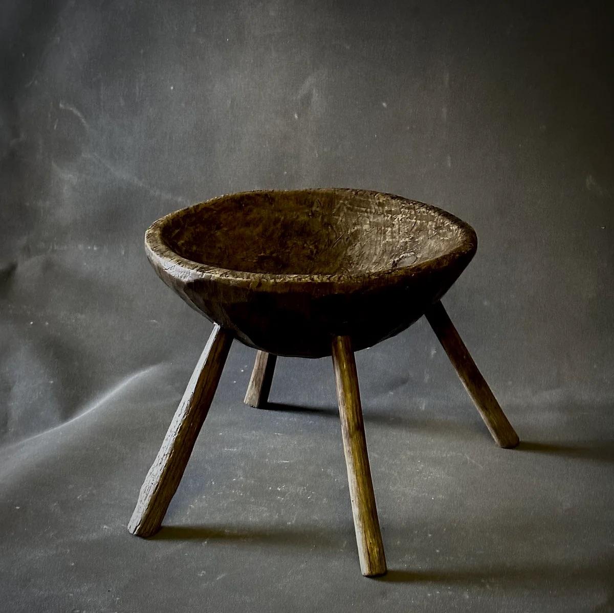 Este primitivo taburete de madera para ordeñar encarna la sencillez rústica con su diseño elemental. El asiento en forma de copa está hecho de una sola pieza de madera, y descansa sobre una robusta base de cuatro patas que se apoya en el suelo. La
