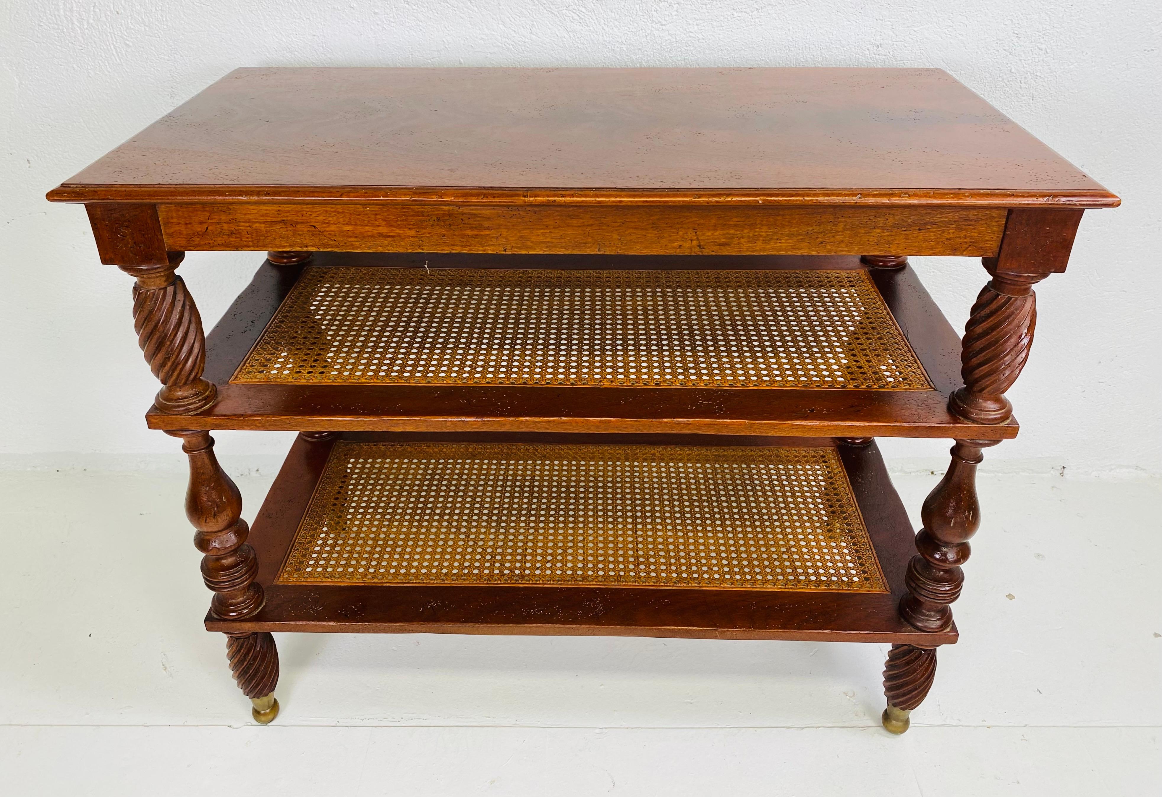 This is a vintage British colonial style mahogany three tiered table. This table has beautifully carved columns and stands on four carved swirled legs with brass details at the bottom. The table features two shelves with caning and the top is a