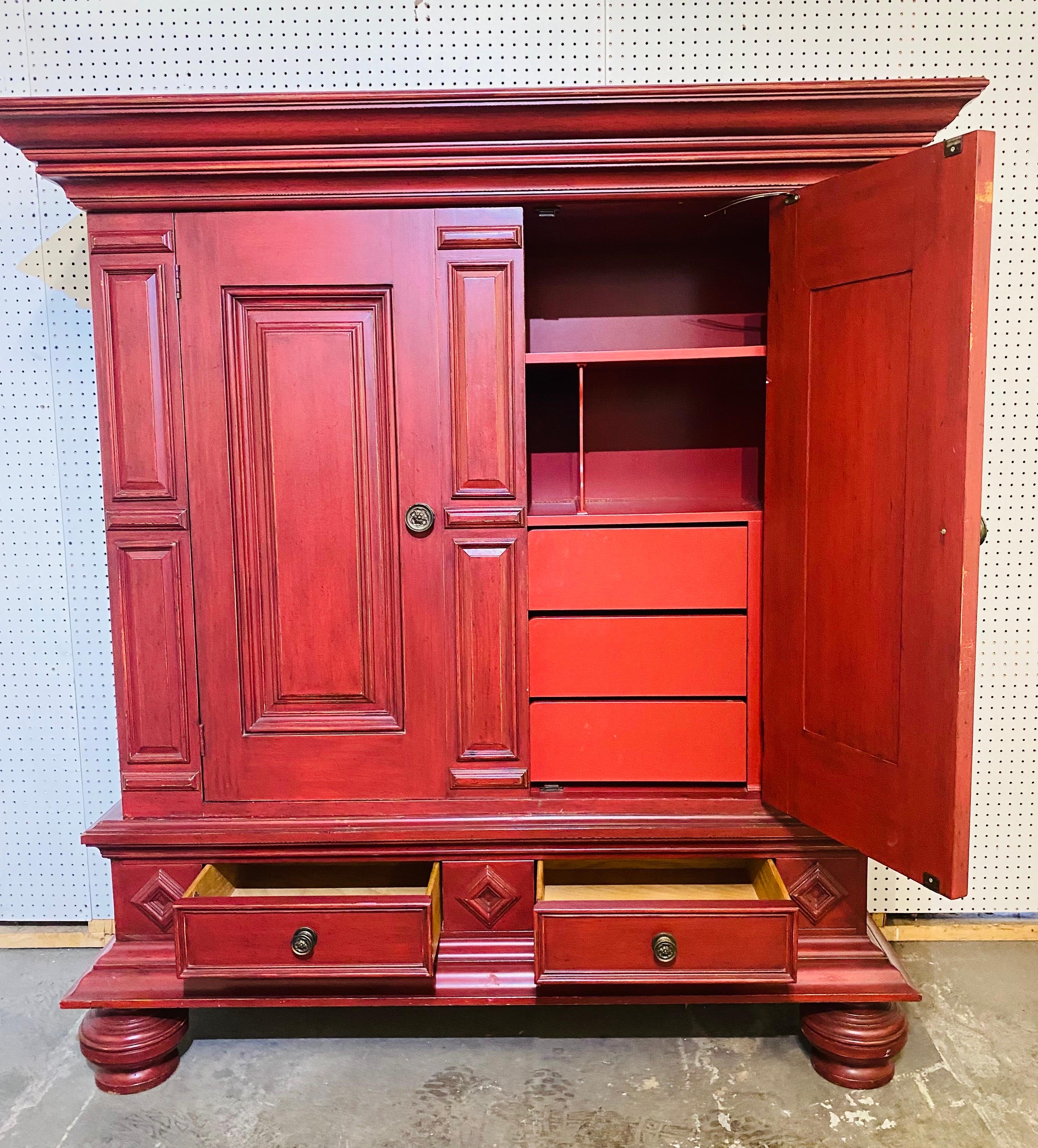 This is a Dutch style two door armoire by milling Road the Baker furniture Company. This two door Dutch style armoire is painted firehouse red and has a custom fitted interior with three drawers and shelving. The armoire stands on four oversized bun