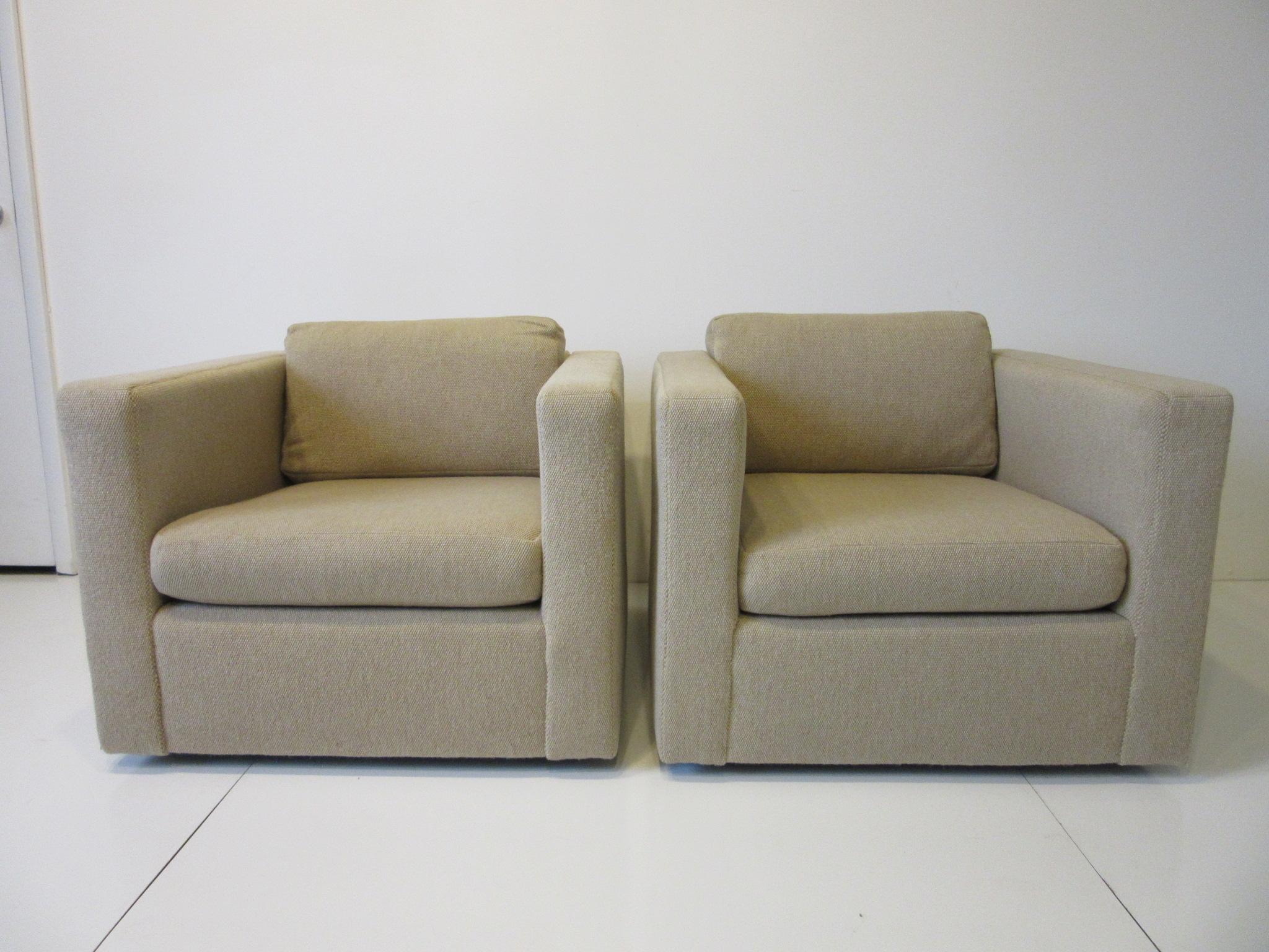 A pair of two cushion cube lounge chairs with tightly woven contract fabric in a sandy beige tone having low wood legs keeping the piece floating off the floor. Retains the manufactures label by the Thayer Coggin furniture company, a simple design