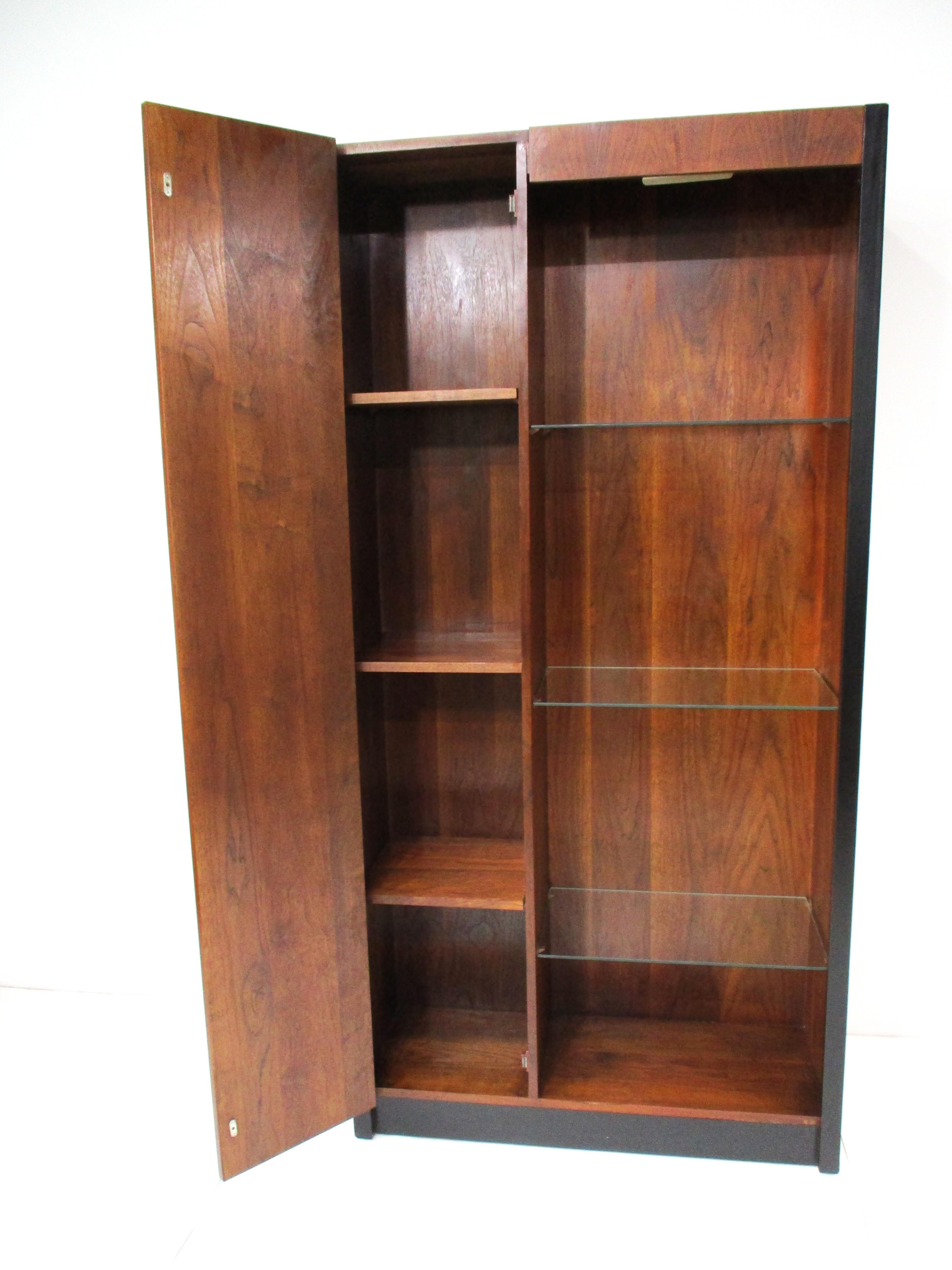 A very well-crafted Mid-Century walnut and satin black painted cabinet bookcase with a door to the left side having three shelves for storage, two that are adjustable. The right side has three adjustable glass shelves and a light to the upper