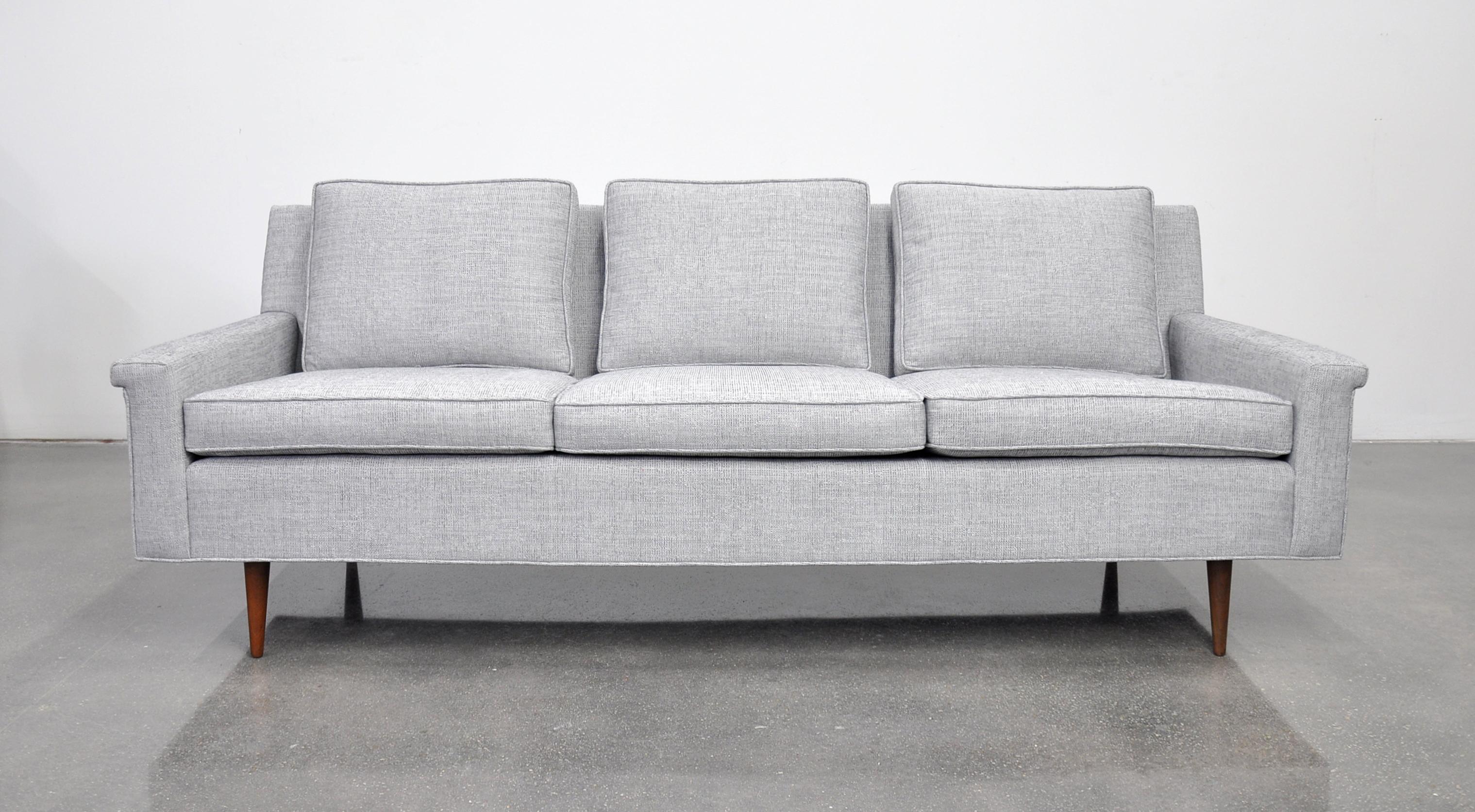 A Mid-Century Modern couch with walnut legs, dating from the 1950s and newly reupholstered in a light grey chenille. The vintage three-seat sofa, designed by Milo Baughman for Thayer Coggin, features loose cushions and round tapered legs. Very