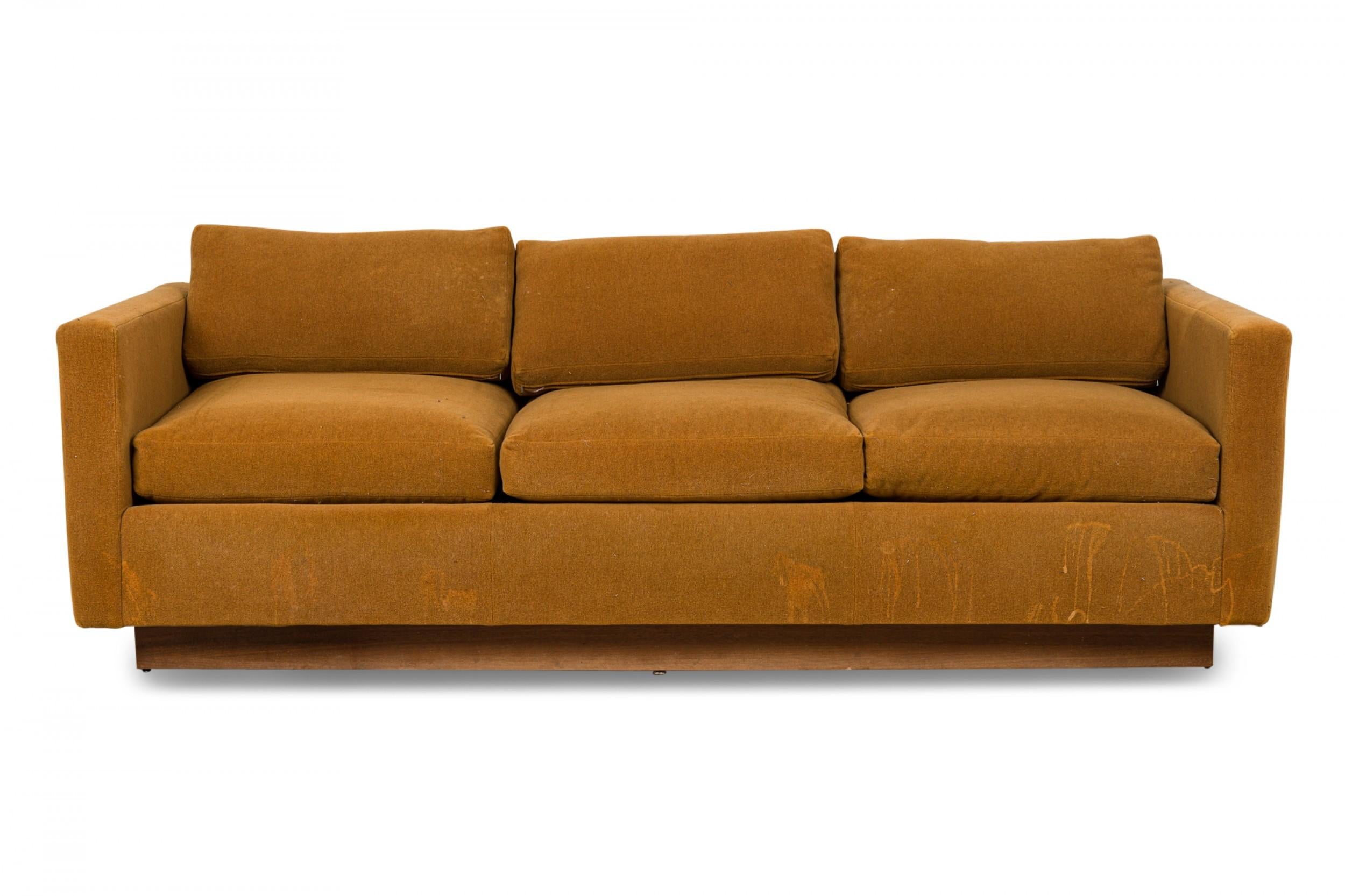 American Mid-Century 'Tuxedo' form sofa in gold colored fabric upholstery resting on a wooden platform base. (MILO BAUGHMAN)
