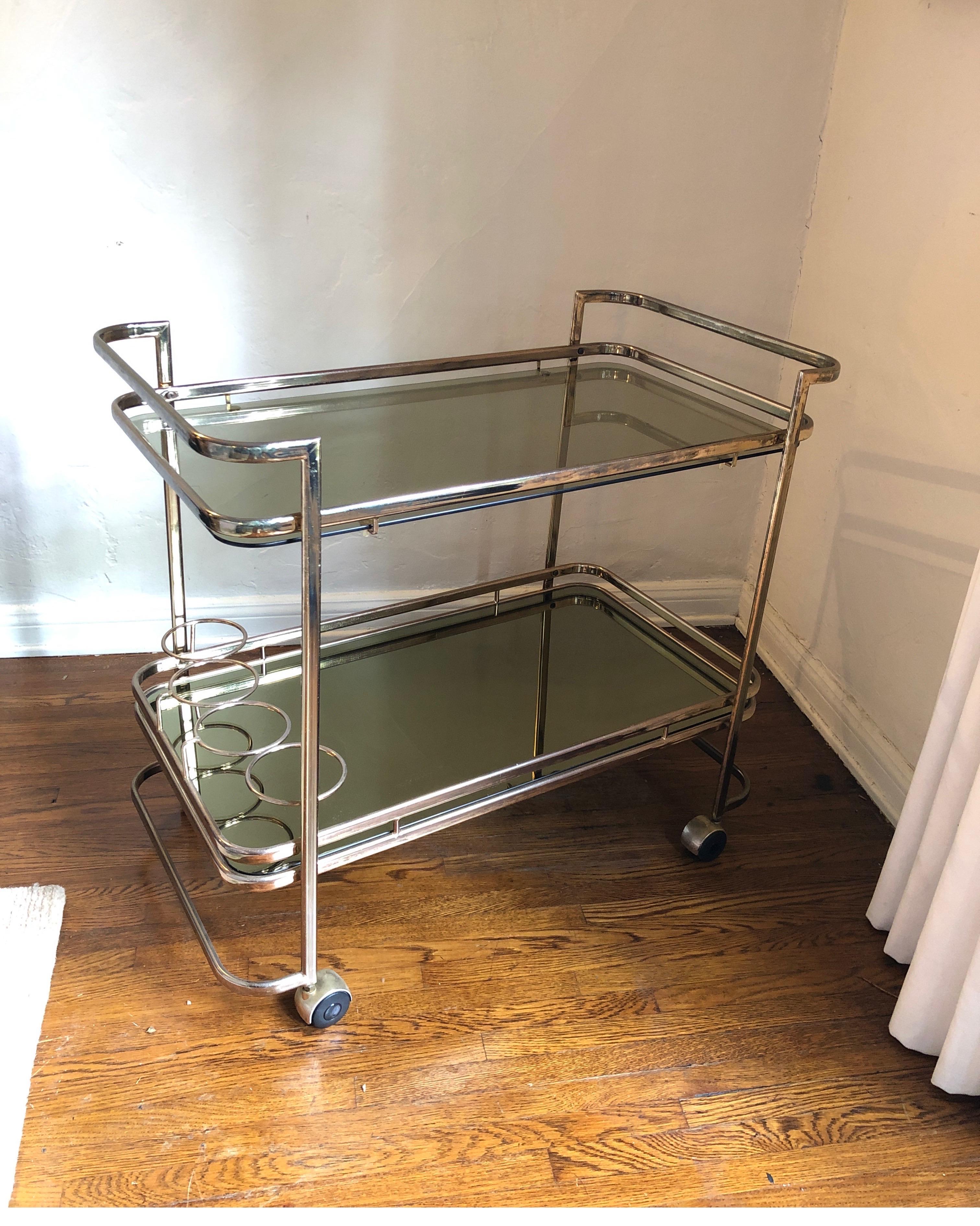 Mid-Century Modern/Hollywood Regency two-tiered brass bar cart with smoked glass top and smoked mirror bottom shelf. 
Sleek Art Deco lines. Glass and mirror both have a gold undertone tint.
Bottom part has holder for 4 bottles/mixers. 

All wheels