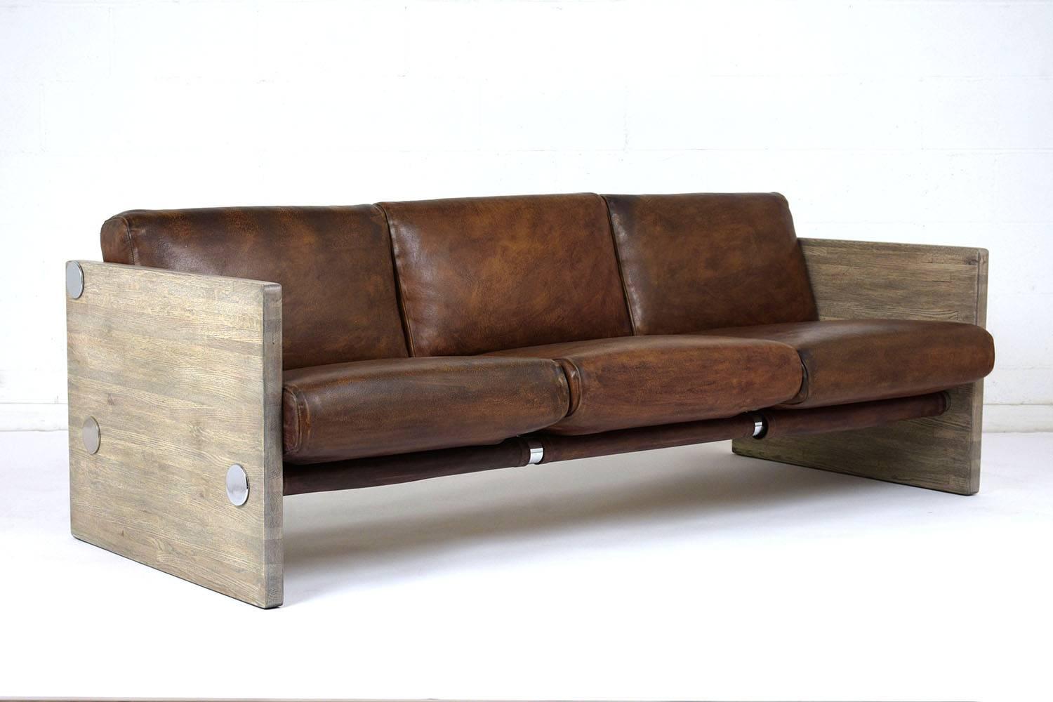 This 1960s Milo Baughman Style Mid-Century Modern Sofa has comfortable leather cushions, a sleek profile, and has been fully restored. The sofa features wood armrests with a faded stacked wood-look finish, steel bar support frame, and the