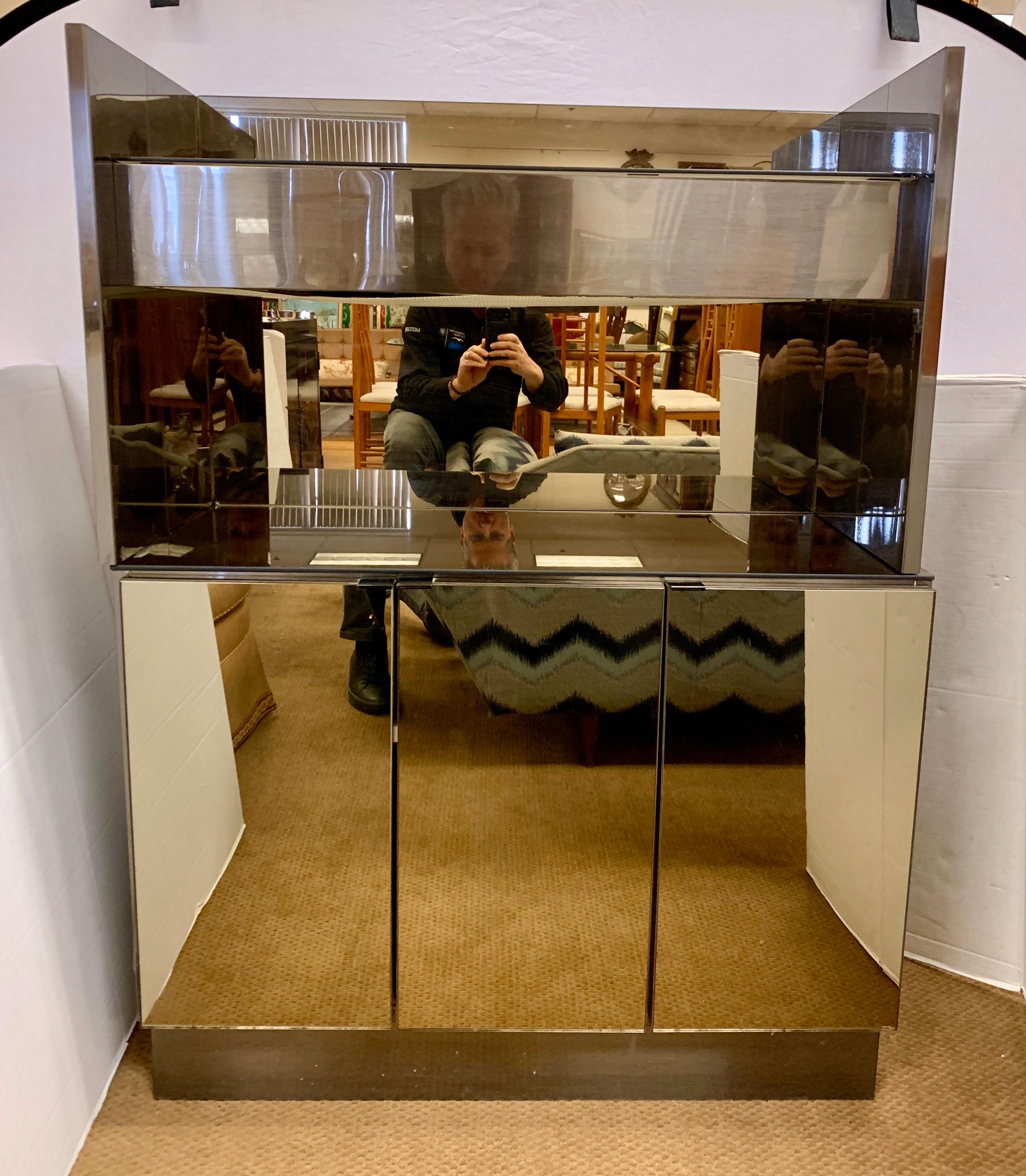 Iconic midcentury two tiered illuminated dry bar where lighting is under top shelf. The cabinet has three doors that open to ample storage ample storage space.. The composition is glass and chrome and the glass is smoked to give it a sleek look. The