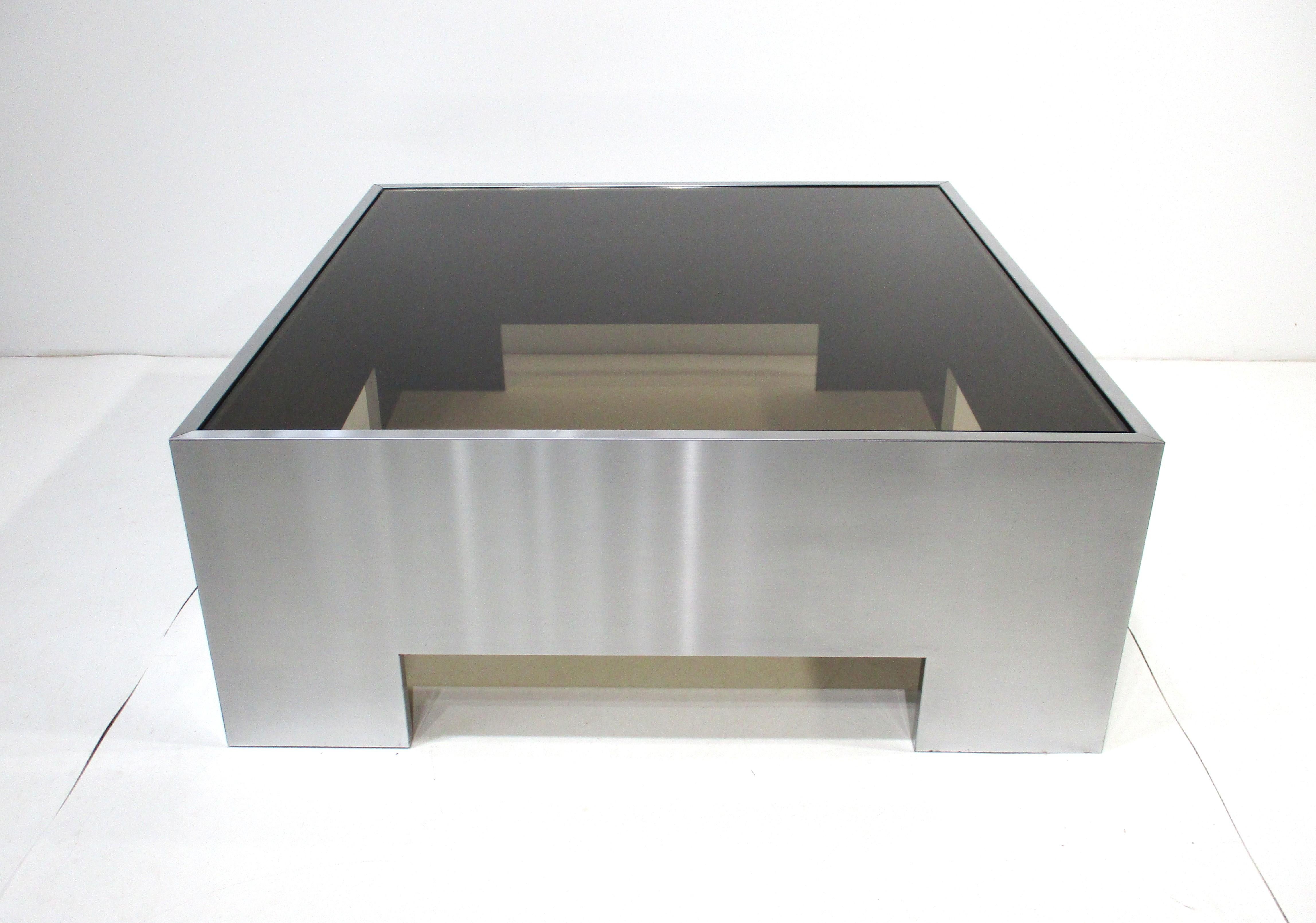 Une table basse carrée sophistiquée en acier inoxydable avec un plateau inséré en verre fumé donnant à la pièce un aspect très épuré. Avec ses extrémités découpées qui donnent à la base de la table un aspect plus léger, cette table basse est une