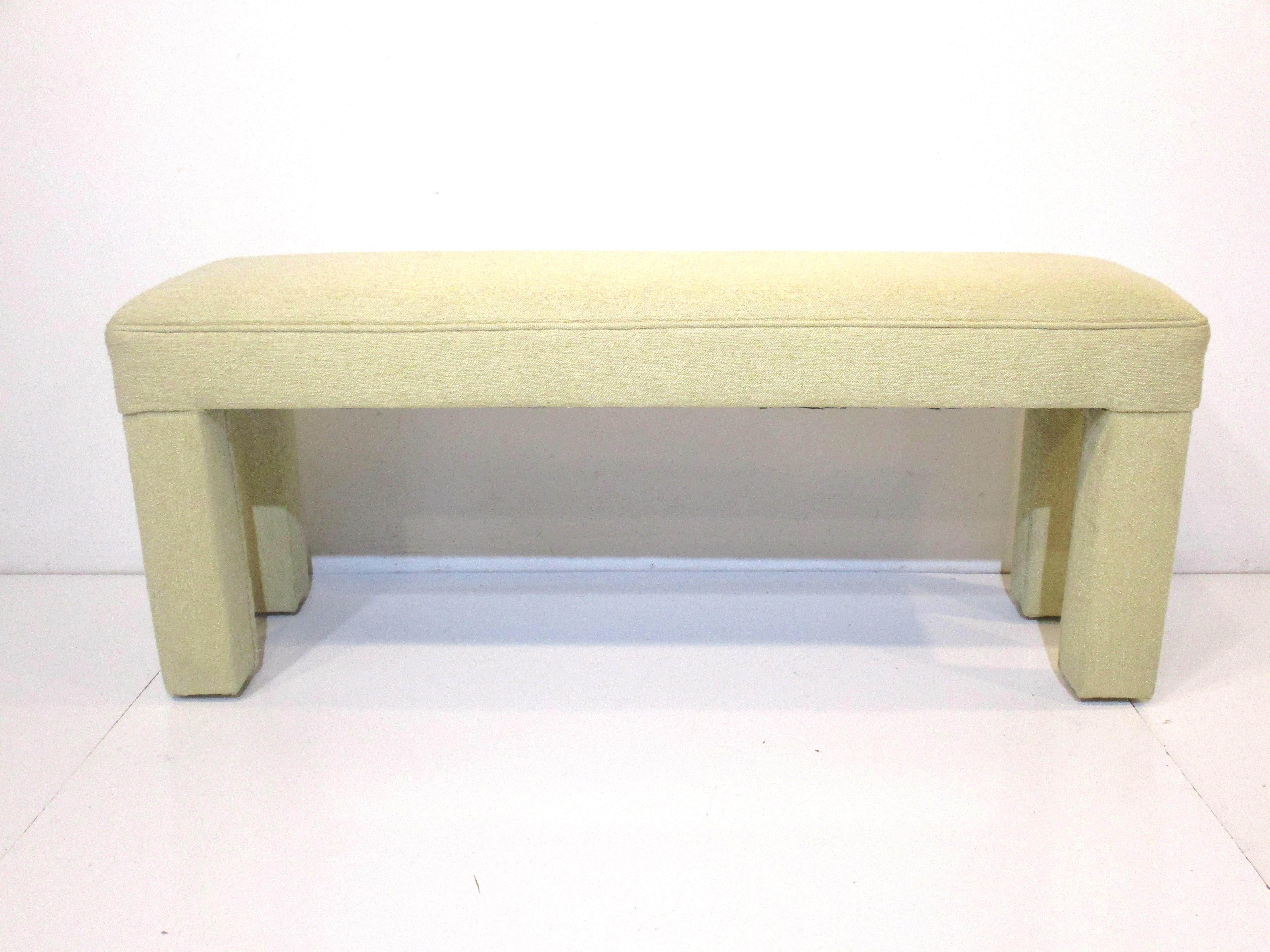 A nice slimer sized bench upholstered in a dark cream nubby contract fabric with covered legs in the style of Milo Baughman . The piece would work perfectly in a tight entrance way because of it's size or at the foot of a bed.