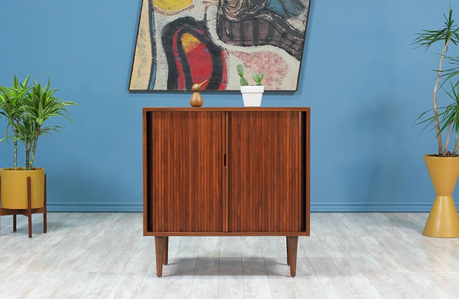 Mid Century Modern Cabinet designed by Milo Baughman for Glenn of California in the United States circa 1950’s. This compact cabinet is made of walnut wood and features two tambour doors with recessed pulls that reveal a shelved interior.
