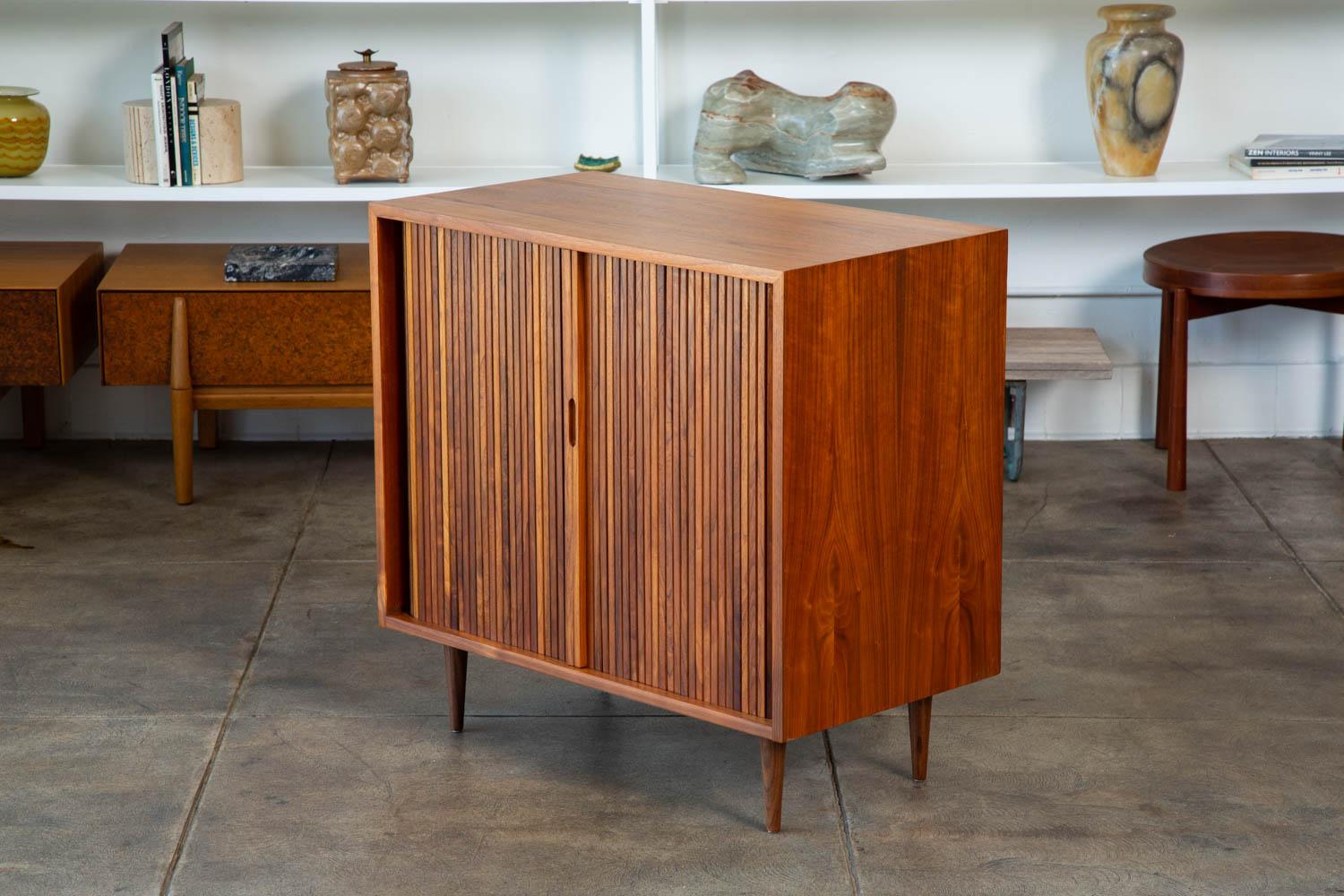 A Milo Baughman for Glenn of California tambour door cabinet. The cabinet features an oiled walnut frame and doors with a sliding walnut shelf atop three red painted drawers. We have two cabinets available and they are priced