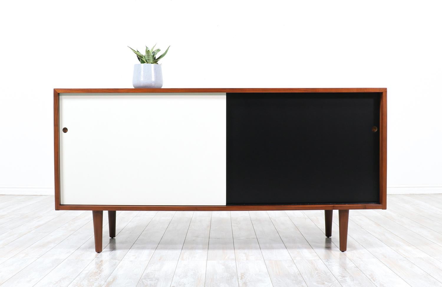 Mid-Century Modern walnut credenza designed by Milo Baughman and manufactured by Glenn of California. circa 1950’s. This compact credenza features two sliding black and white doors with brass pulls that open to reveal a shelved compartment on each