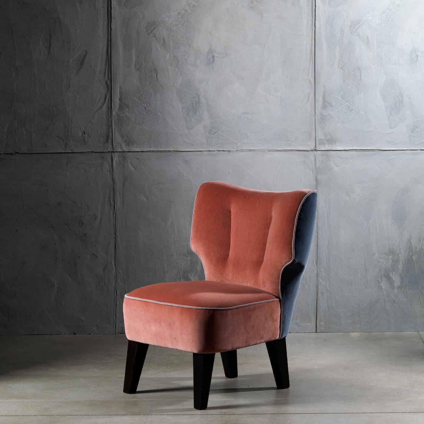 This low-sitting, cozy armchair features a roomy, spacious velvet seat and a back in the soft, comforting colors of powder pink and blue, creating a silhouette defined by surprising, harmonious volumes. It stands upon wooden legs with a dark walnut