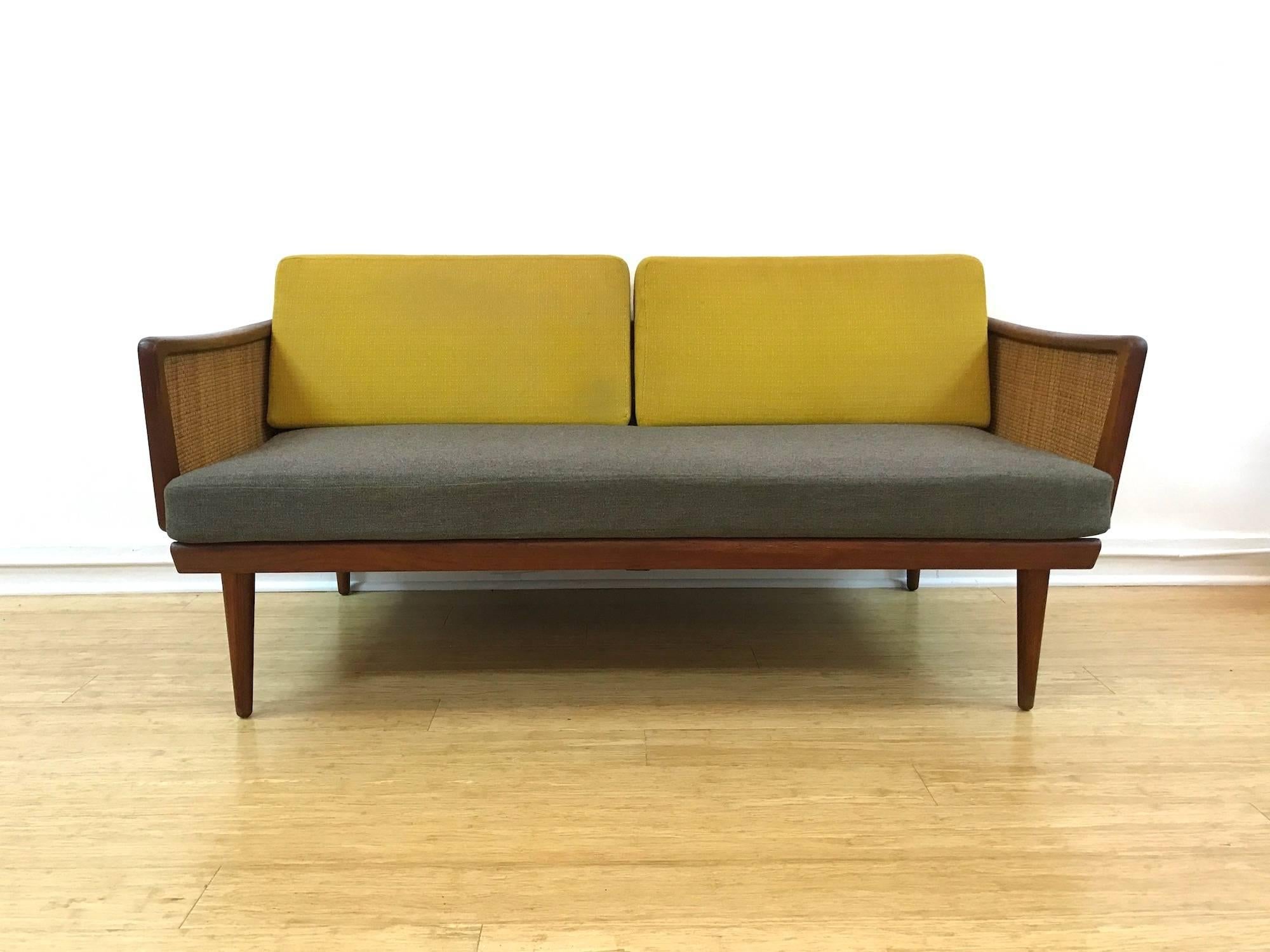 Minerva Danish Modern loveseat daybed designed by Peter Hvidt & Orla Mølgaard-Nielsen in teak and cane for France & Sons, Denmark, c. 1960, retailed by John Stuart. 
Shaped arms over caned sides that fold down, original back and seat cushions and