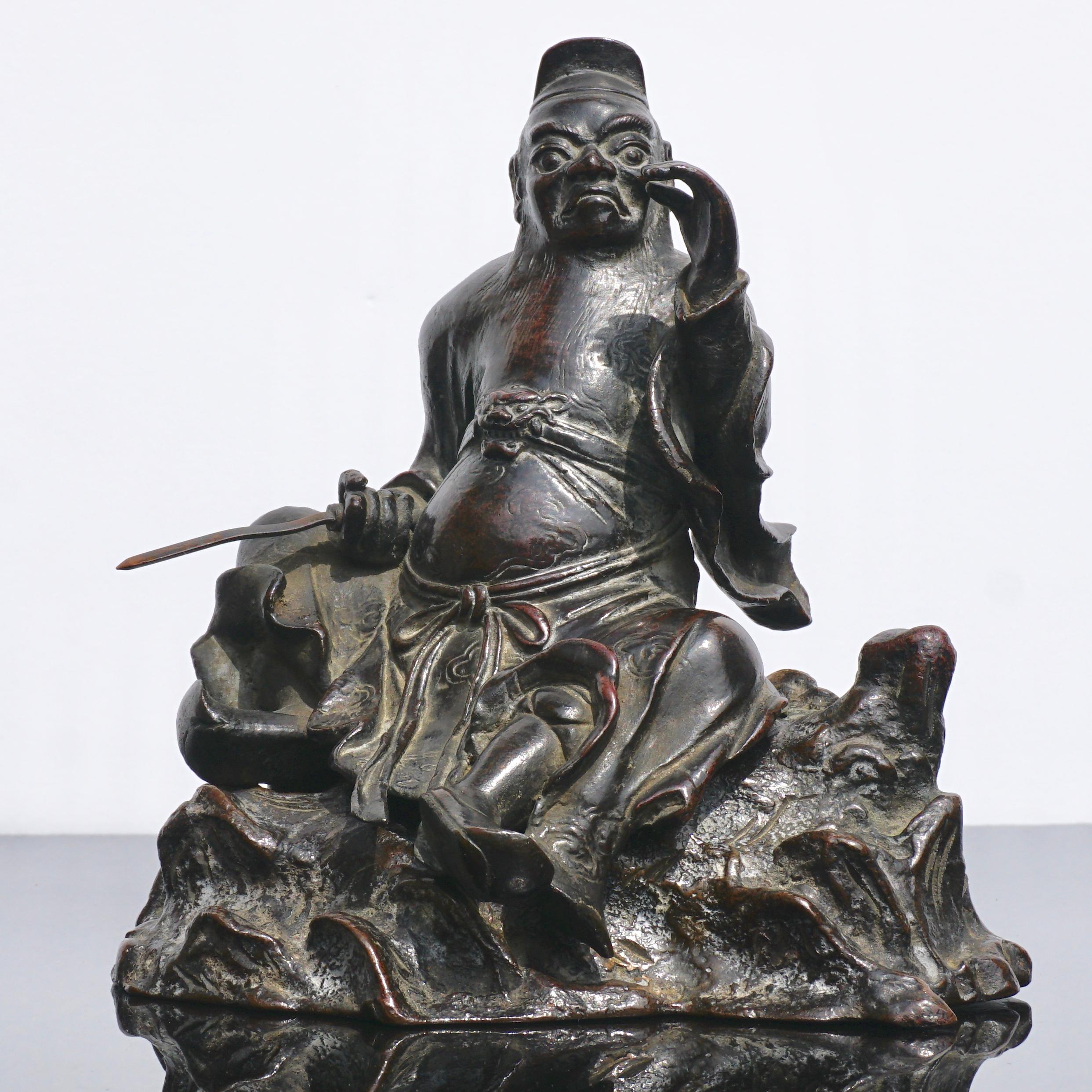 A Chinese Ming dynasty two-piece bronze incense censer or cache in the Form of Guandi or Guan Yu holding a sword in his right hand sitting on a natural boulder. The sculpture or figure with a hat, long beard is elegantly robed with a lion or tiger