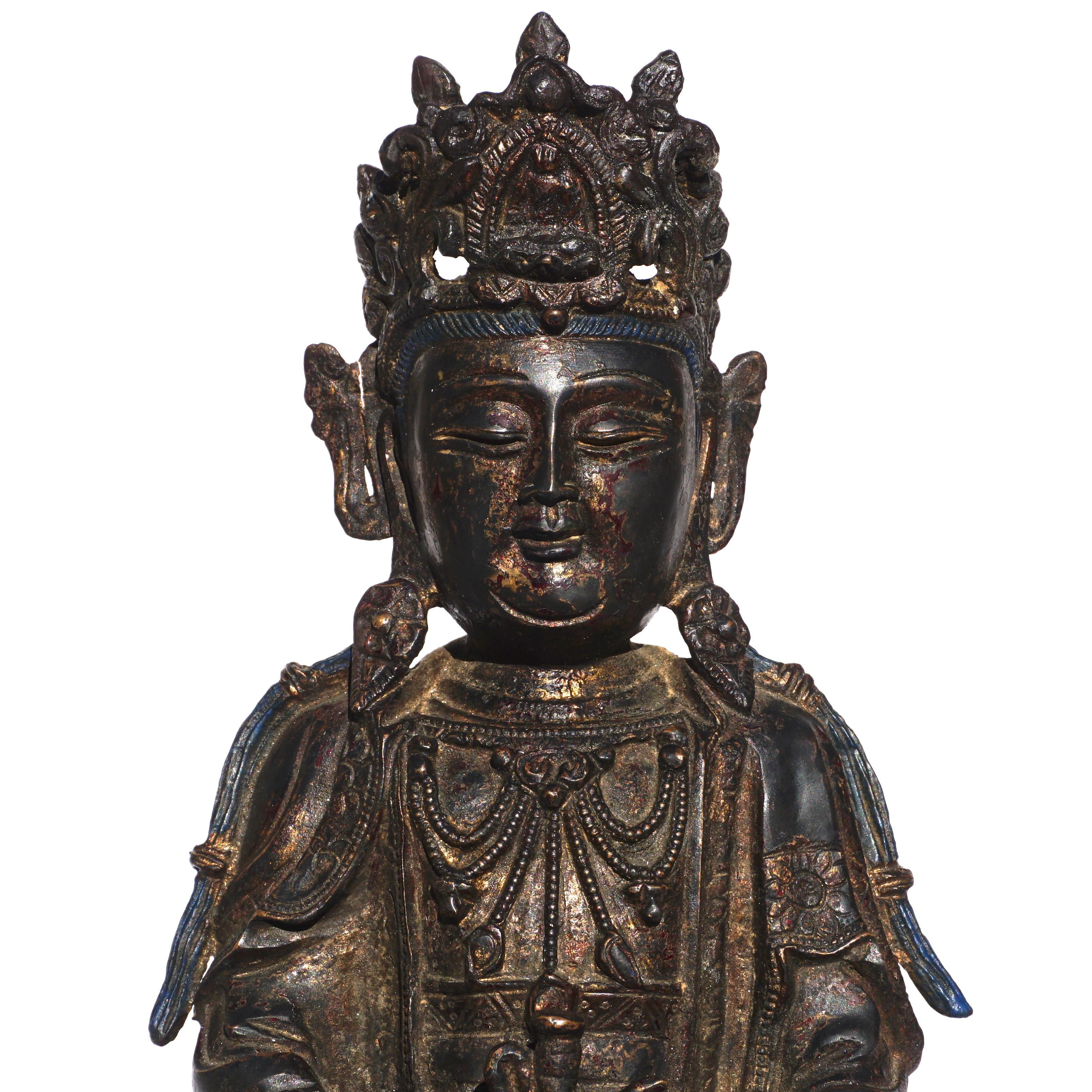 Eine vergoldete und lackierte Bronzefigur der Guanyin aus der Ming-Dynastie. Die Göttin der Barmherzigkeit ist im Dhyanasana sitzend dargestellt:: die Hände in den Schoß gelegt und einen Krug haltend. Ihr langes Gewand ist an der Brust geöffnet und