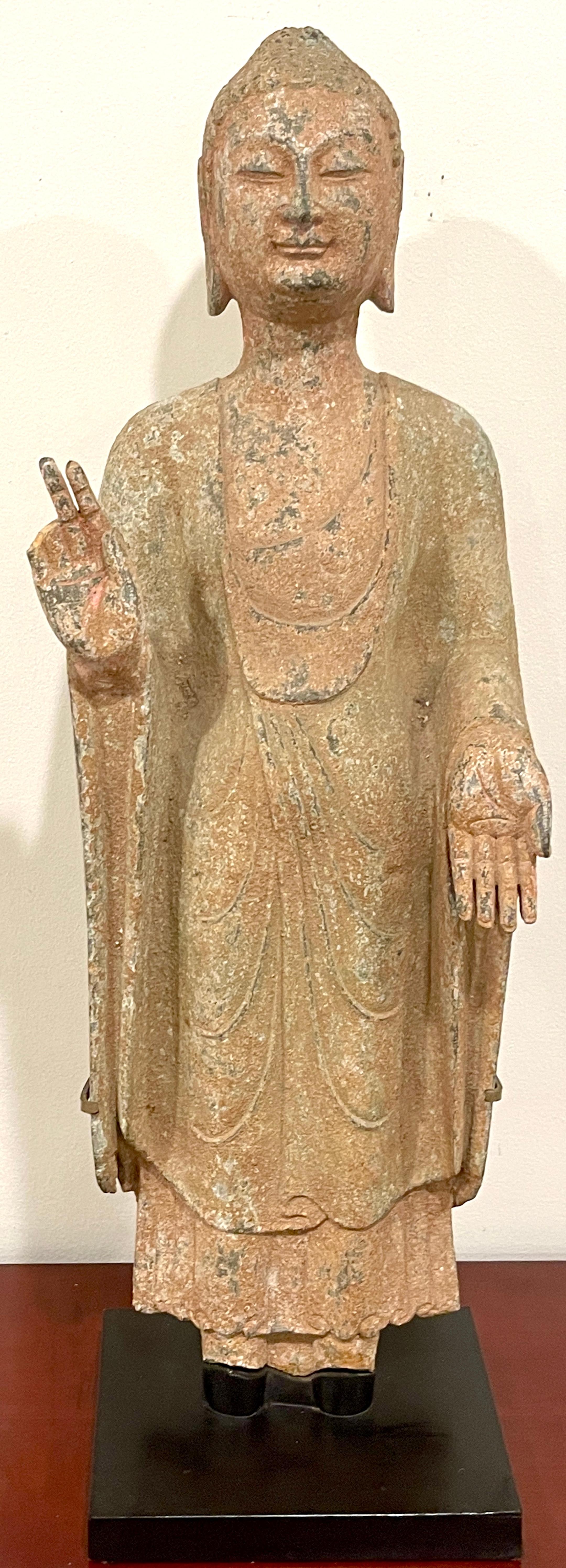 Ming Dynasty Polychrome Grey Stone Standing Buddha, Museum Mounted (1368-1644) 

A polychrome gray stone sculpture of Buddha stands, wearing layered robes that cover most of the body, leaving only a part of the chest uncovered. The left arm is