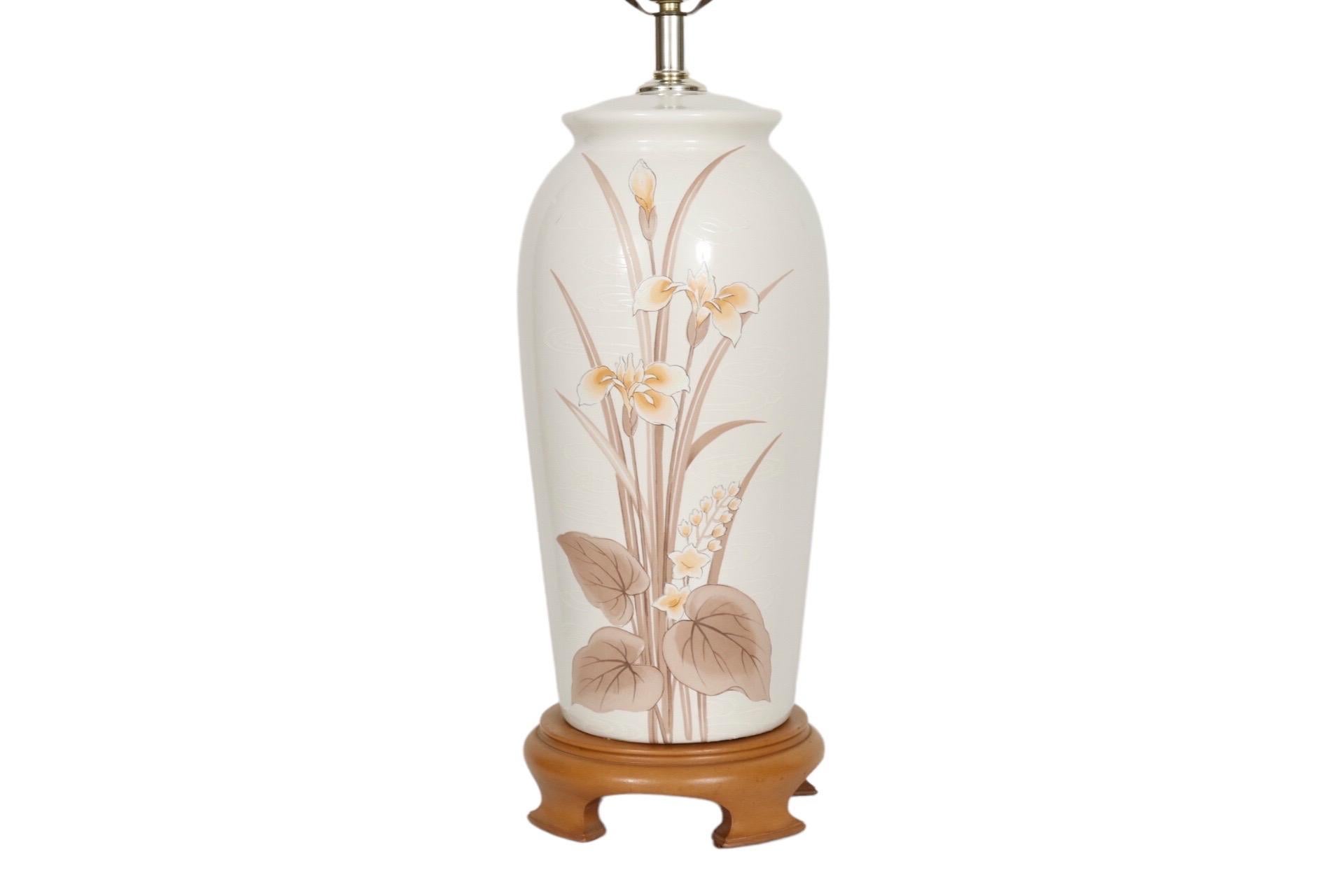 A Ming style ceramic table lamp in white. Decorated with hand painted iris and lily flowers with foliage in a golden brown color palette. Stands on a brown wooden base with four Ming style feet. Measures 23”H to the top of the socket, 33.25”H to the