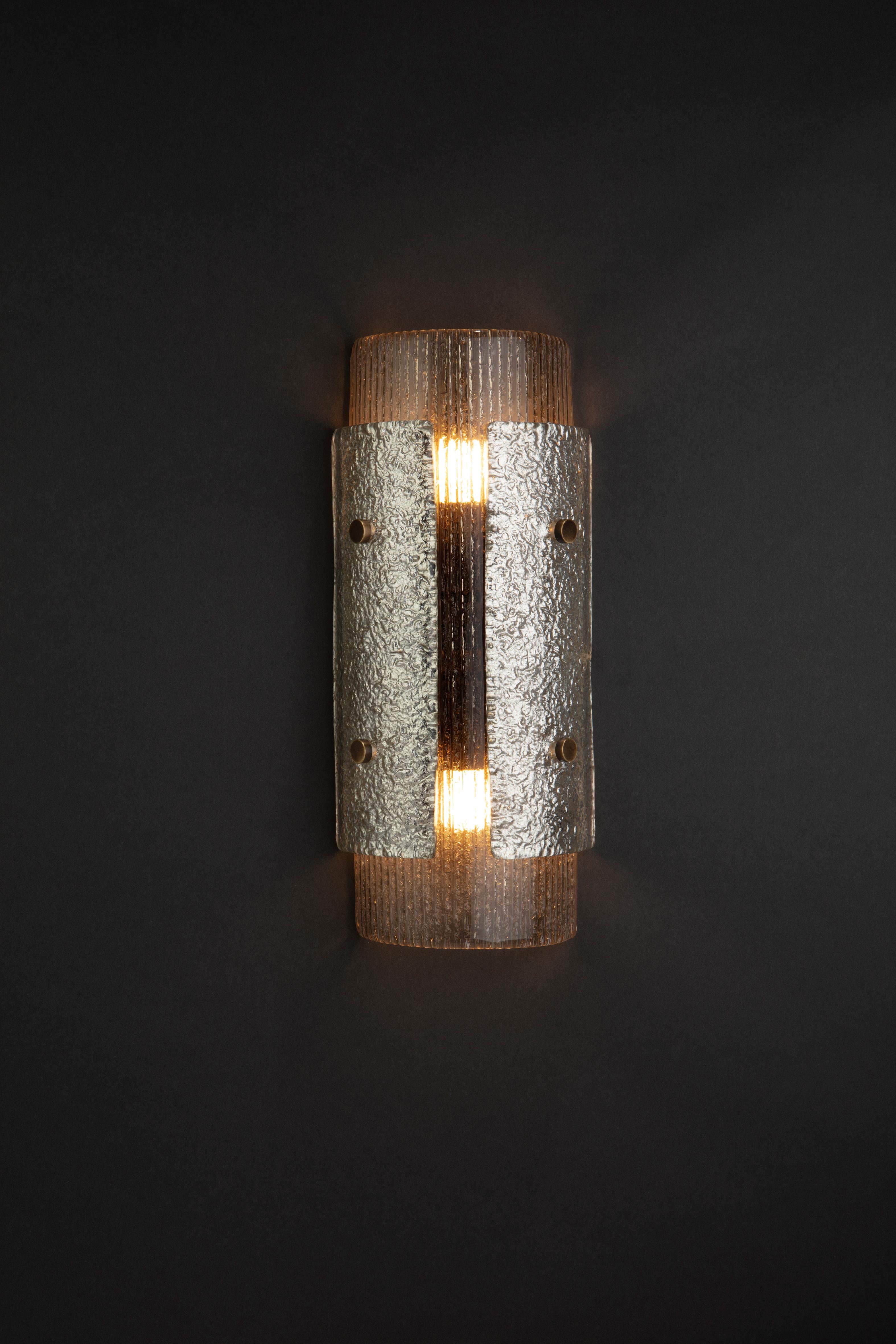 The mini laterali wall sconce is a smaller version of the laterali sconce and with different textures of glass. The body is ribbed which diffuses the light without alternating the colour. The two appliqués have silver gilding on the
