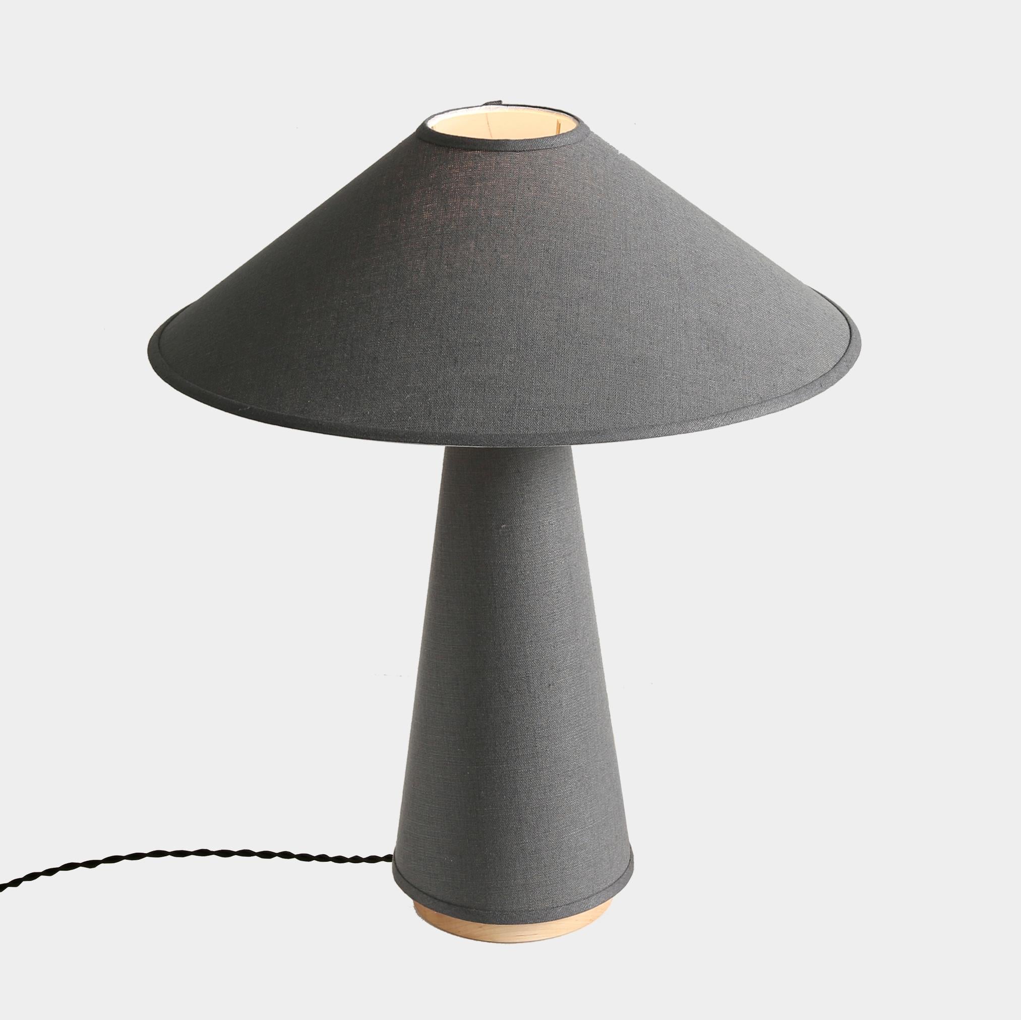 The Linden table lamp features a charcoal grey linen shade and lamp body, solid hardwood maple base, and brass details. The tabletop fixture references the classic profiles and angles of mid-century modern design with distinct clean lines, smooth