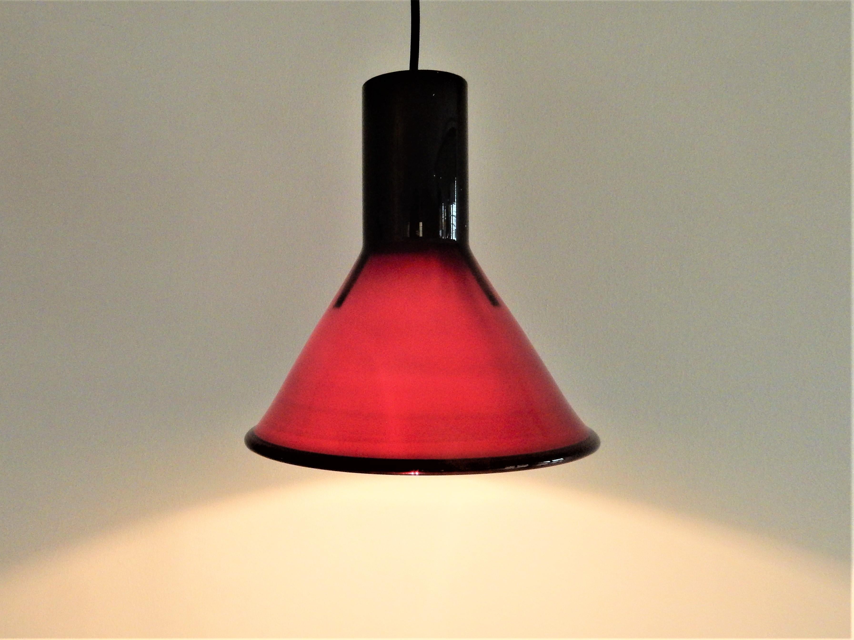 This mini P&T pendant lamp was designed by Michael Bang for Holmegaard in the 1970s. This Danish lamp is made of opaline glass and is black/aubergine on the outside and white on the inside. When lit, the lamp gives a partial warm red light. It is in