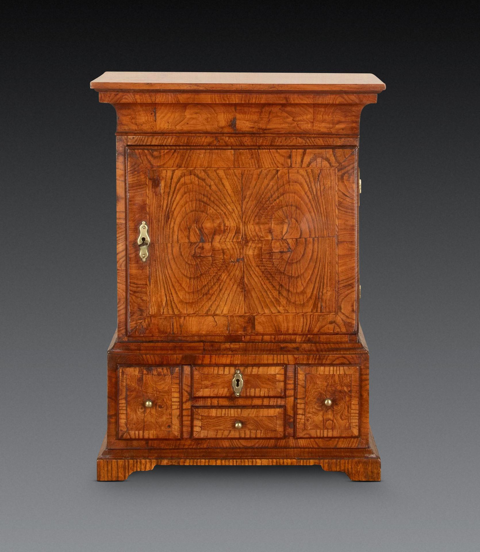 A rare mid C18th miniature table/source cabinet in figured Ash, with swept cornice above a door enclosing an oak arrangement of seven drawers and central arched recess, the base with one drawer simulating four smaller drawer front for aesthetic