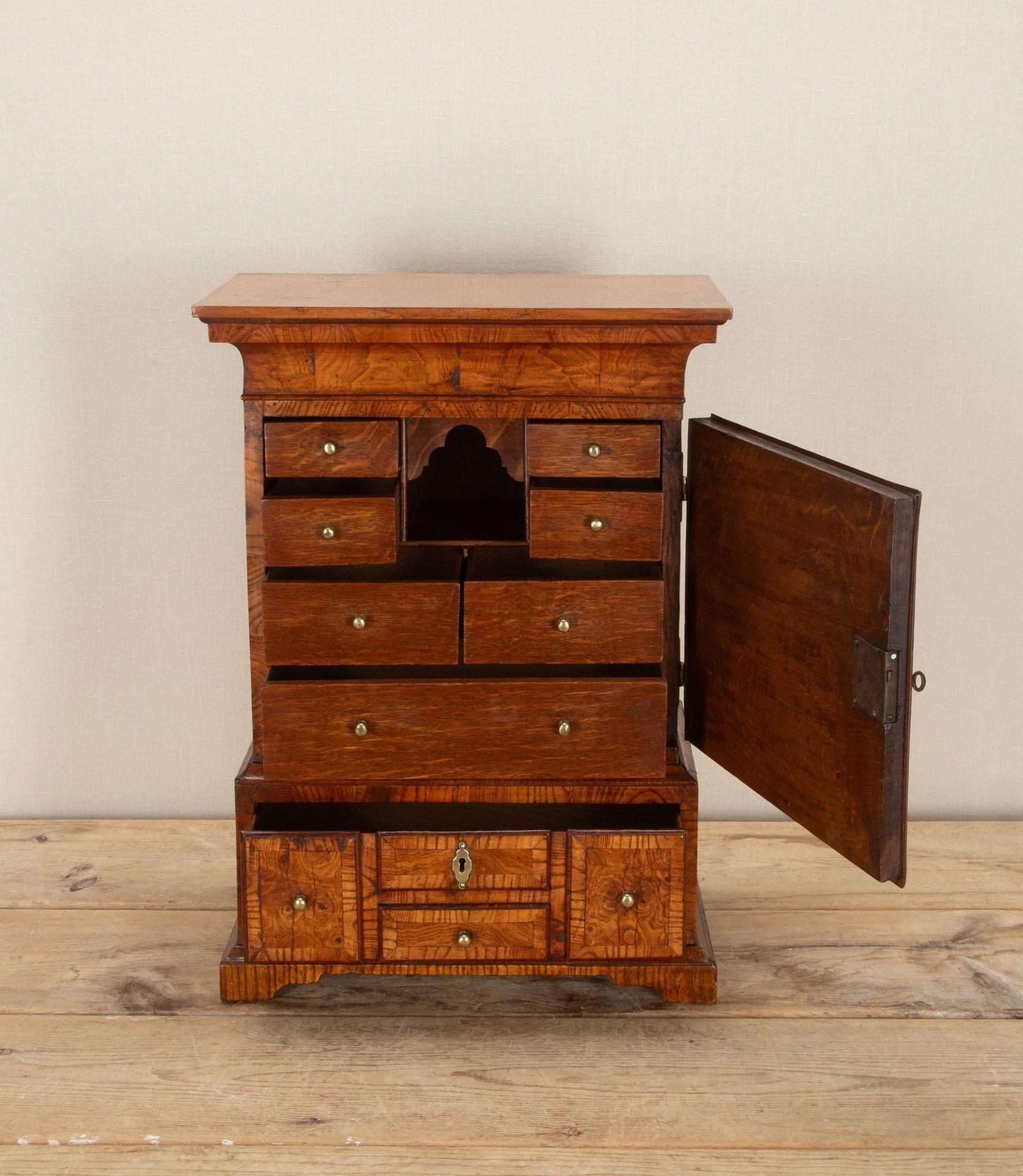 Raro tavolino o mobile in miniatura inglese della metà del XVIII secolo in frassino figurato. Con una cornice a raggiera sopra un'anta che racchiude una composizione di sette cassetti in Oak e una nicchia centrale ad arco. La base è dotata di un