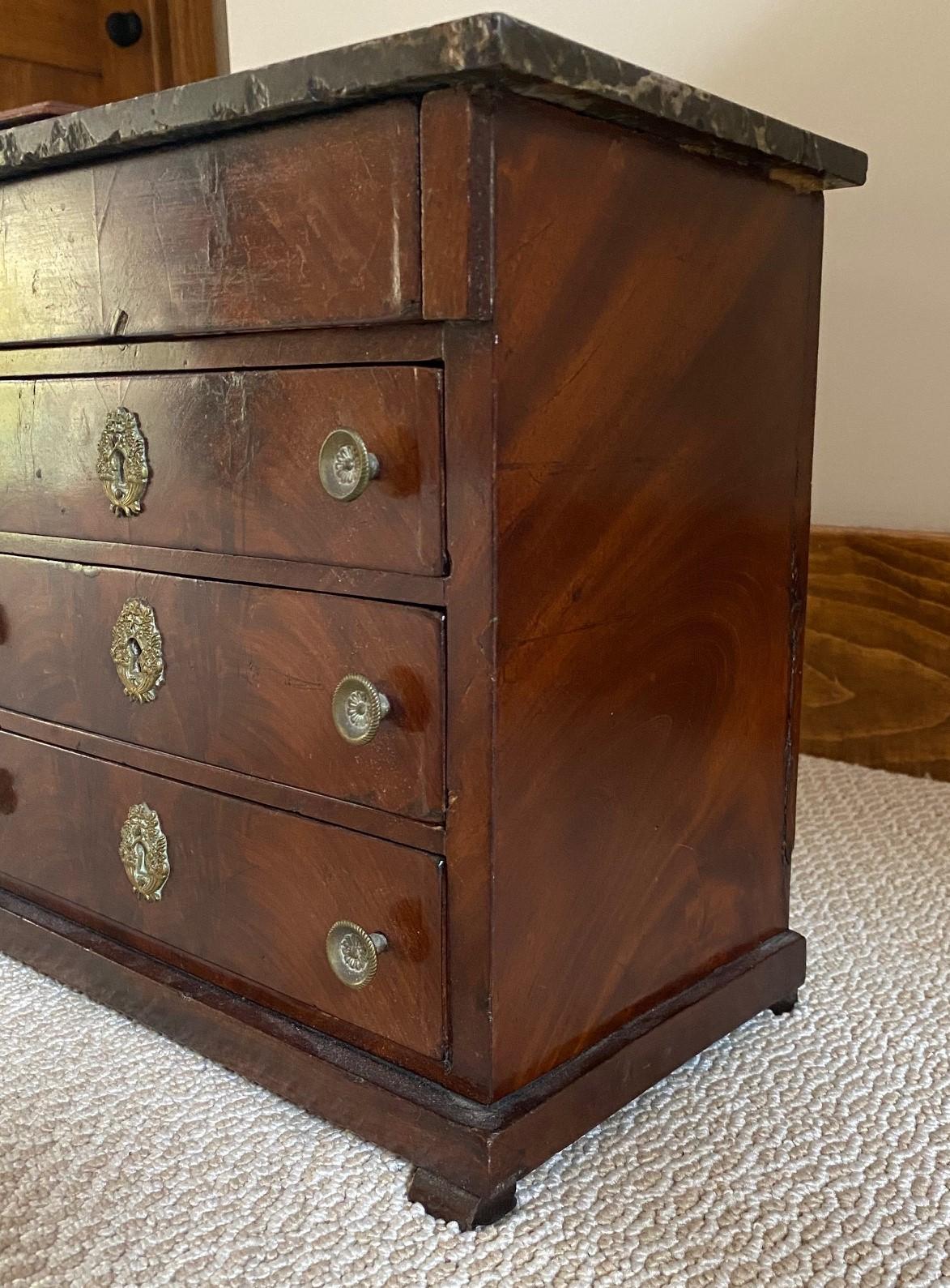 19th c. Charming antique miniature chest of drawers. The chest has a marble top. The case is figured mahogany, with four stacked drawers, the bottom three mounted with brass pulls and escutcheon.

Dimensions: 11.5