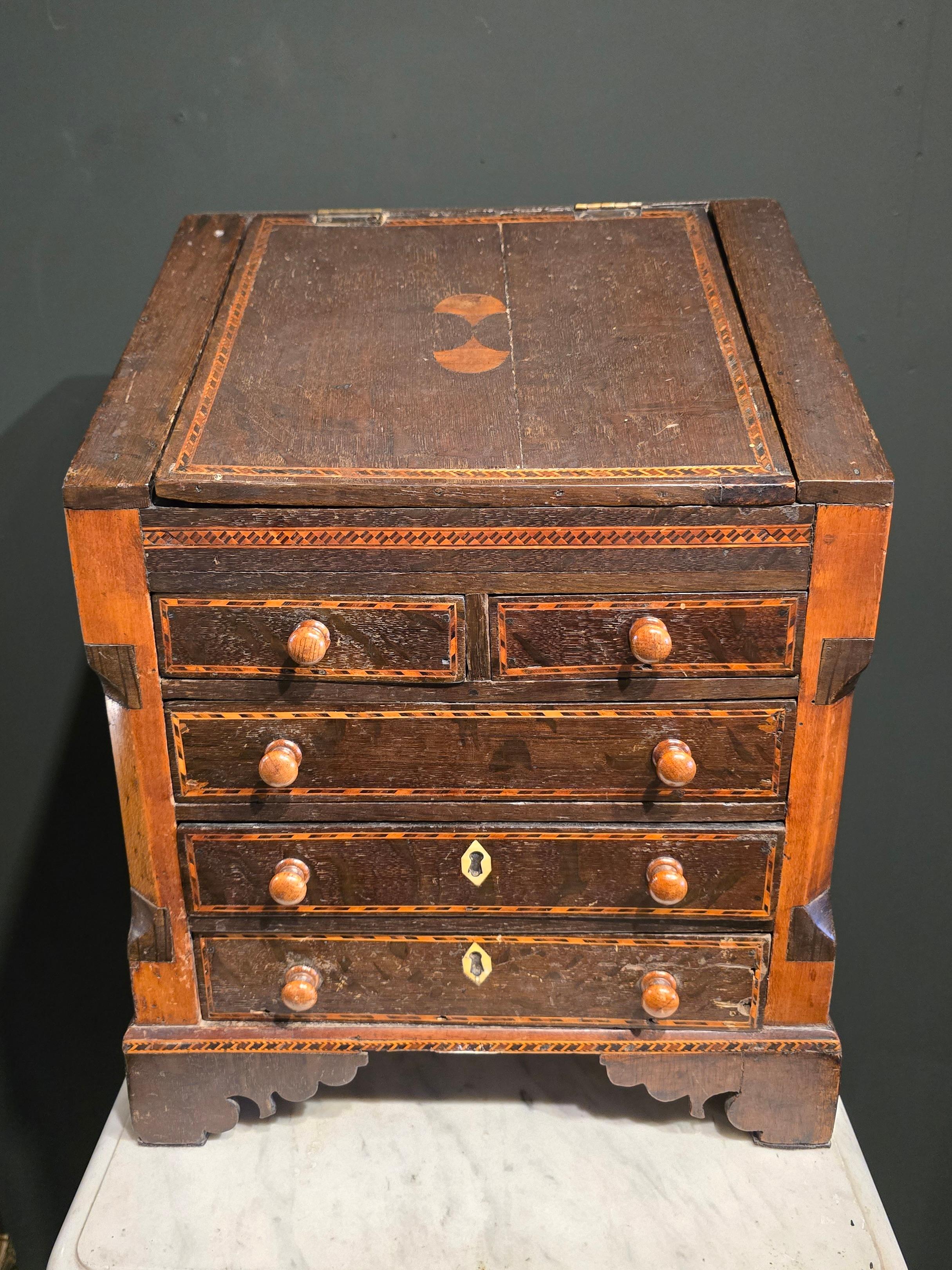 Scrivania antica in miniatura risalente al 1800.  Spesso si parla di campioni commerciali o di pezzi di apprendisti.  Il corpo principale è in Oak, con vari legni intarsiati per la decorazione, Sycamore, Holly, Walnut.  Il cassetto inferiore è