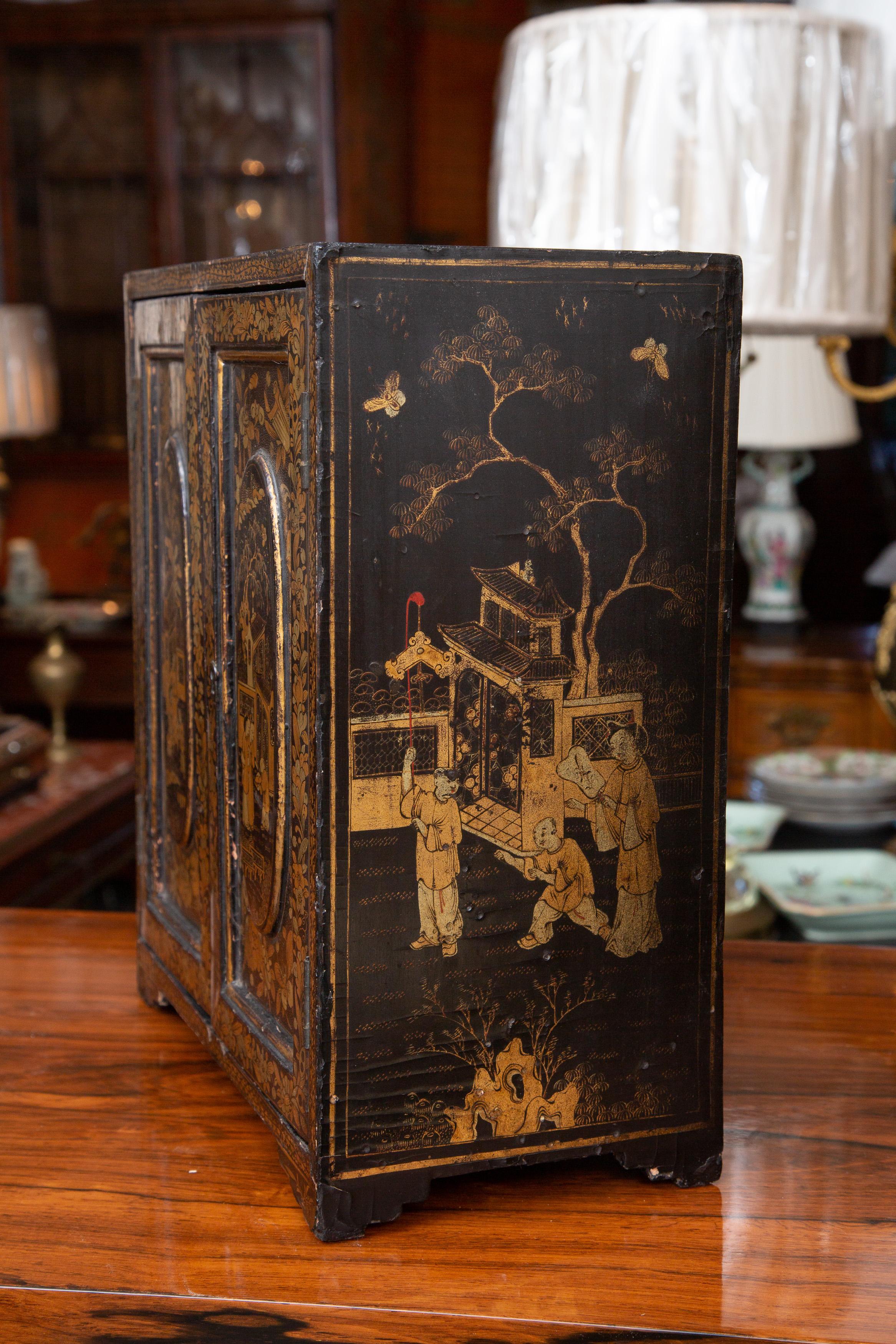 This is a very sophisticated English black lacquered miniature table cabinet painted overall with gilt chinoiseries depicting Chinese village life and foliage. When the cabinet doors are opened, a bank of five small drawers are revealed, circa late