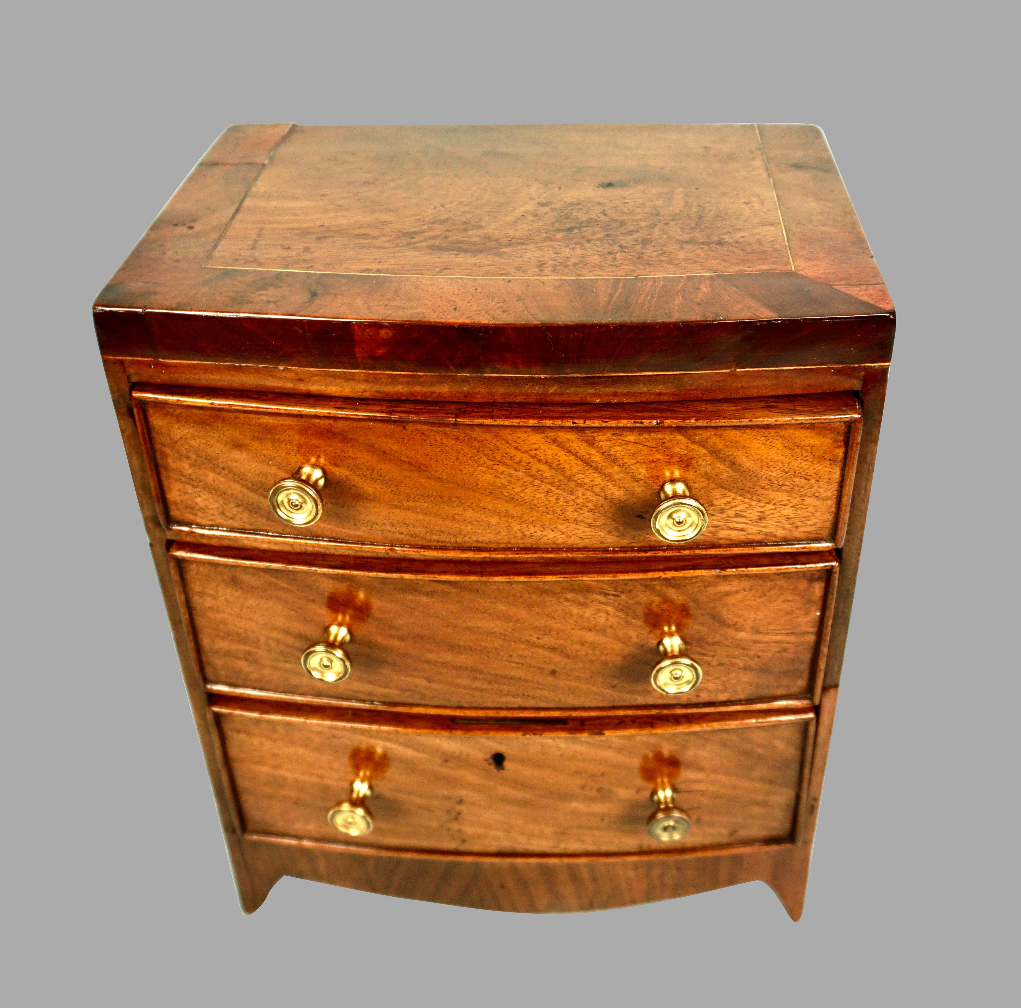 A charming miniature Hepplewhite style 3 drawer chest, the crossbanded top over 3 graduated small drawers resting on French feet. This miniature is very true to form of full-sized chests of this style. Now with replaced brass knobs, this piece would