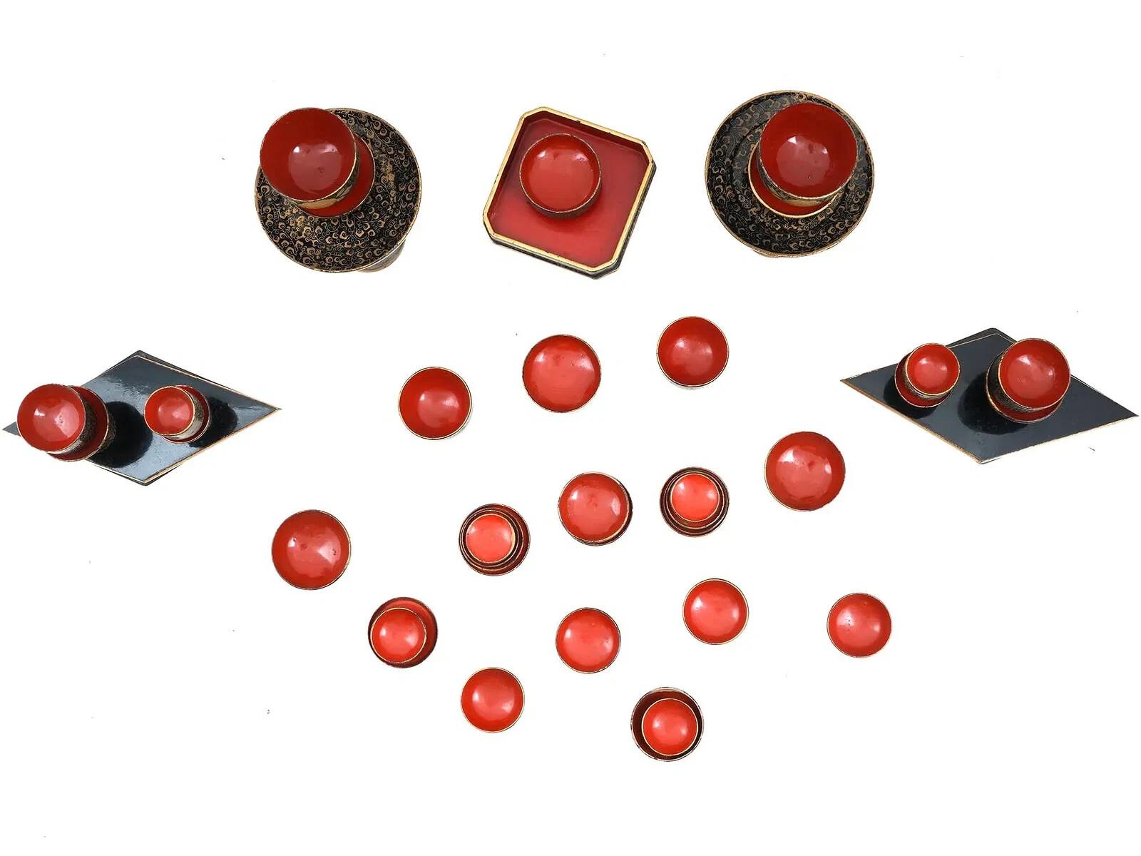 A miniature Japanese set made of wood and lacquered in a classic black with gold accents and lacquered red interiors. The set consists of a variety of small bowls, cups, and stands, each with a delicate design of gold foliage pattern. There are two