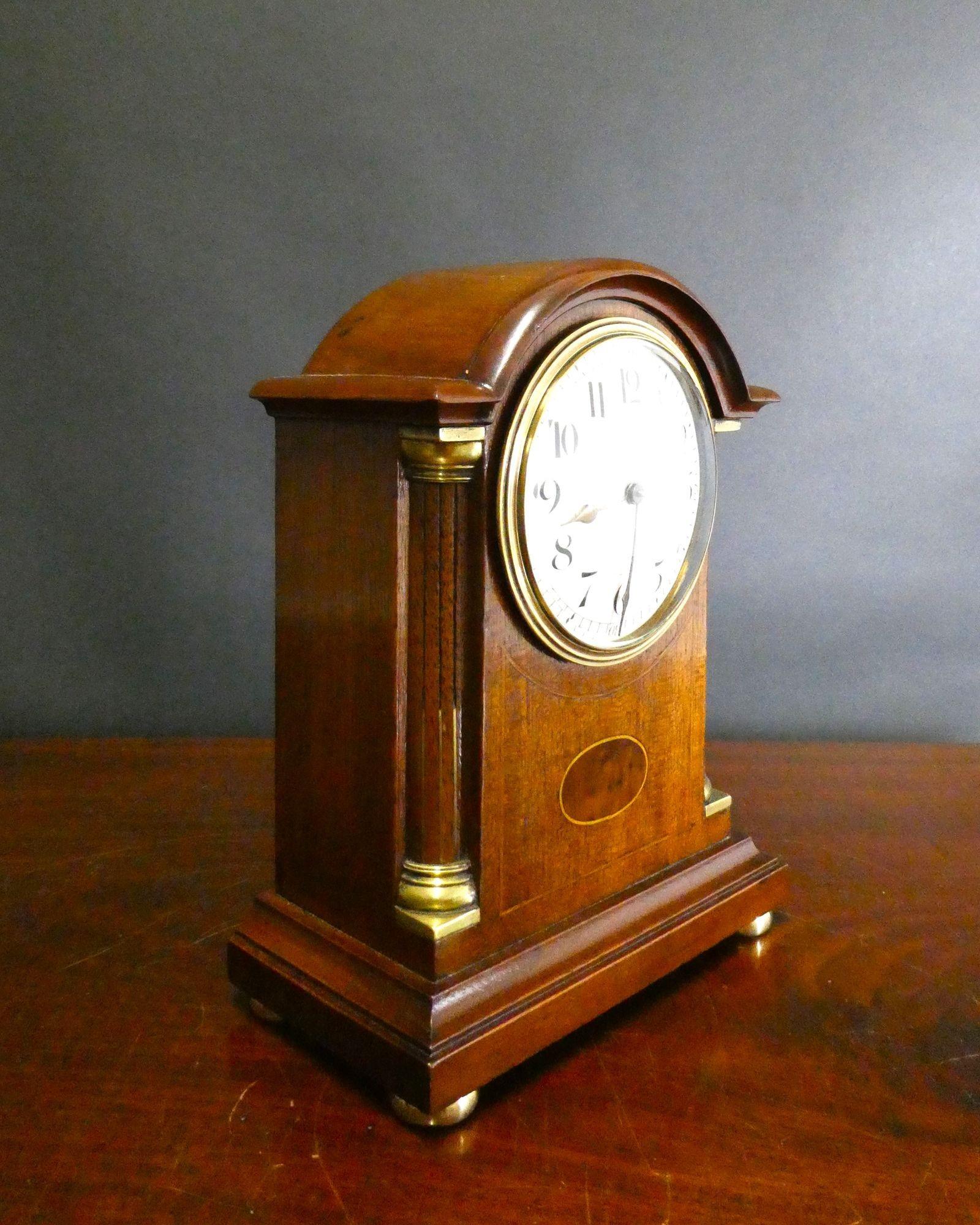 Miniature mahogany mantel clock housed in a break arch case with reeded pillars and brass capitals. Inlaid central cartouche with line inlay and resting on a stepped, moulded plinth with four brass bun feet.
Enamel dial with Arabic numerals,