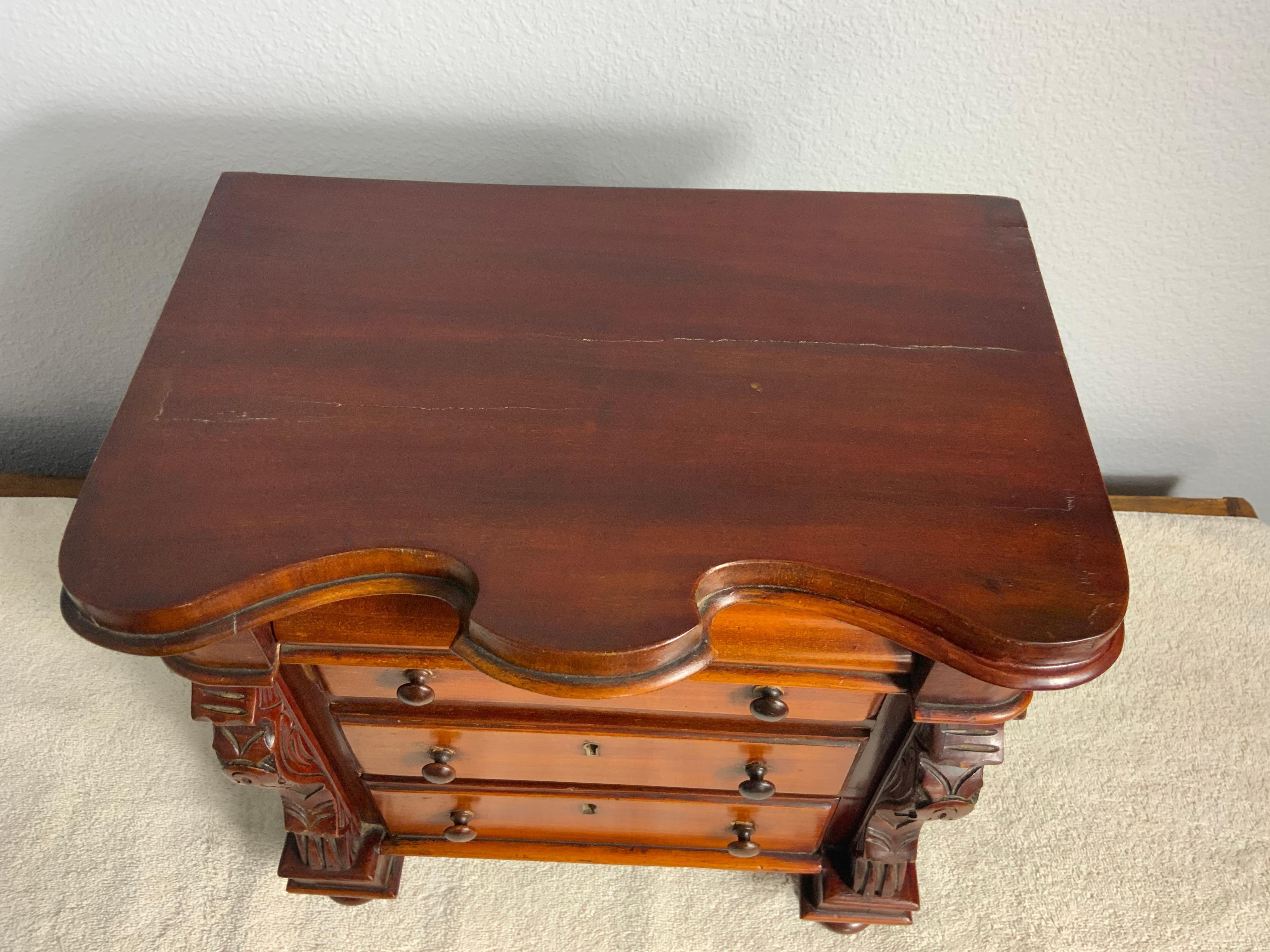 Una cassapanca in miniatura in mogano di epoca vittoriana, dal 1850 al 1870. Due o tre cassetti graduati con incastro a coda di rondine a mano, centrati tra due colonne laterali intagliate. I tre cassetti inferiori hanno tiranti e serrature