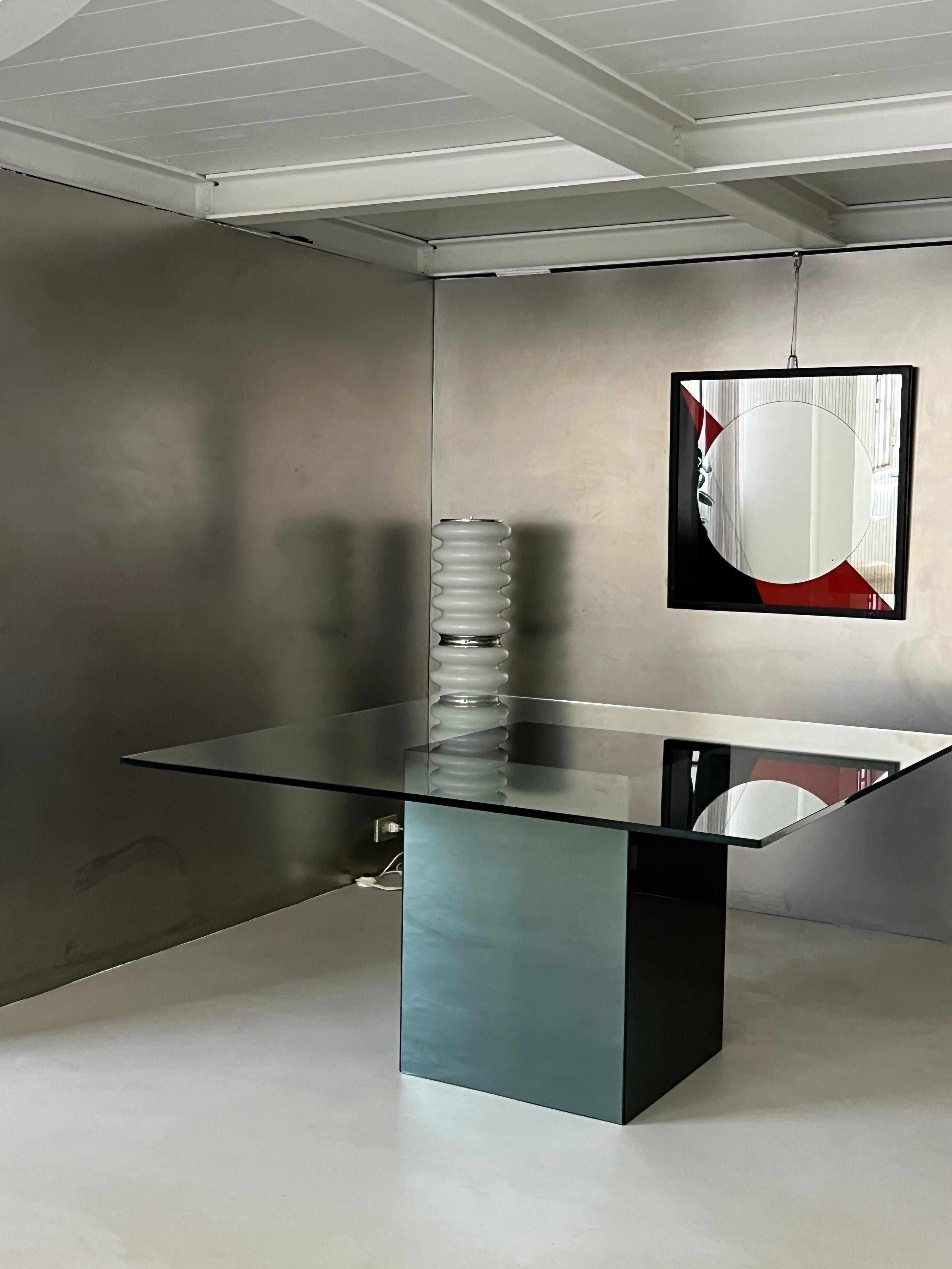 The vintage square table designed by Nanda Vigo features a sleek and modern aesthetic characteristic of her minimalist style. It has a square-shaped design with a sturdy central base, which is covered in smoky mirror, creating a reflective, slightly