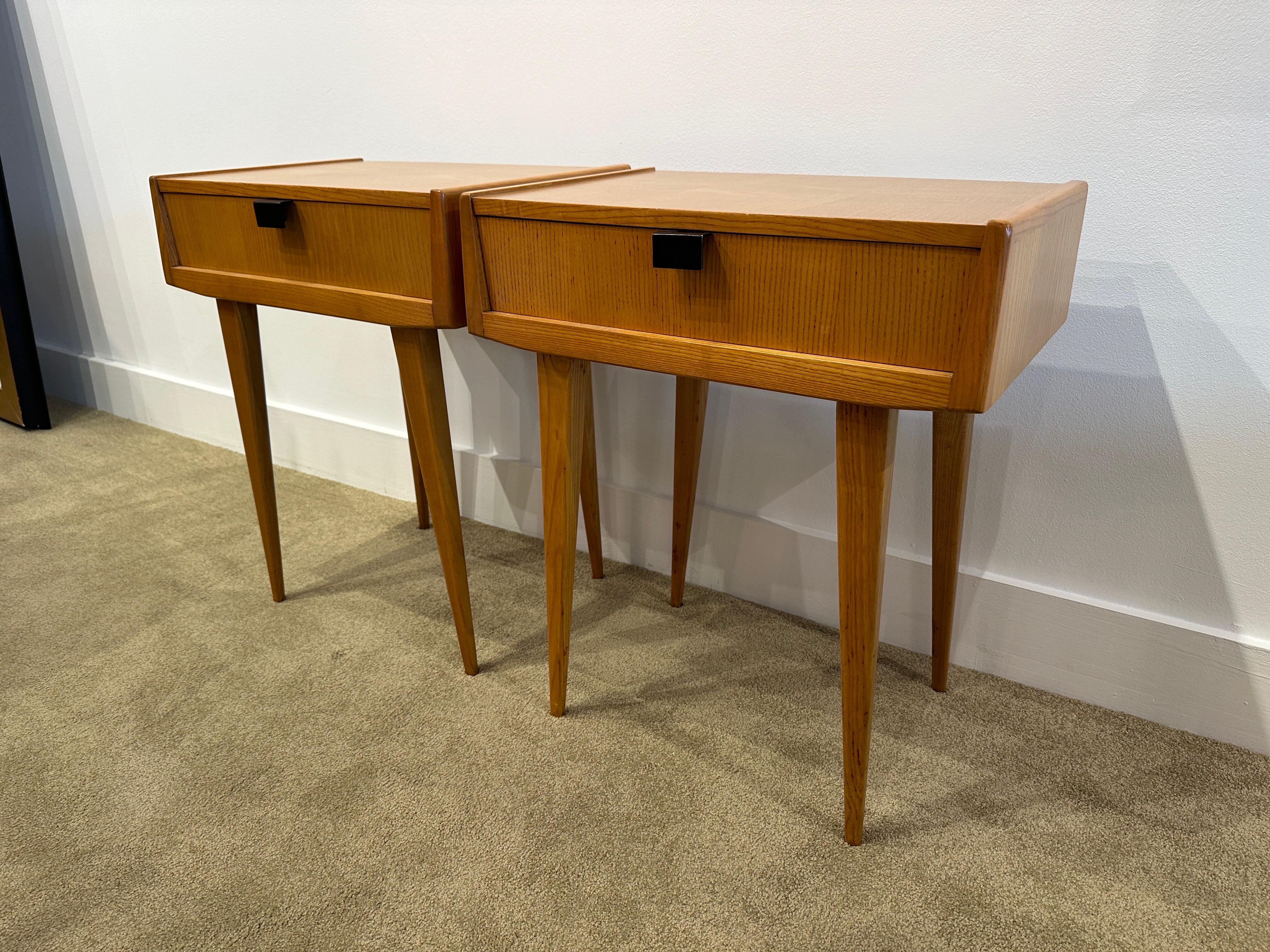 Extremely handsome pair of side tables/ nightstands with clean lines, elegant elongated legs and rich oak veneer. Original metal pulls and very sleek.  THIS ITEM IS LOCATED AND WILL SHIP FROM OUR EAST HAMPTON, NY SHOWROOM.