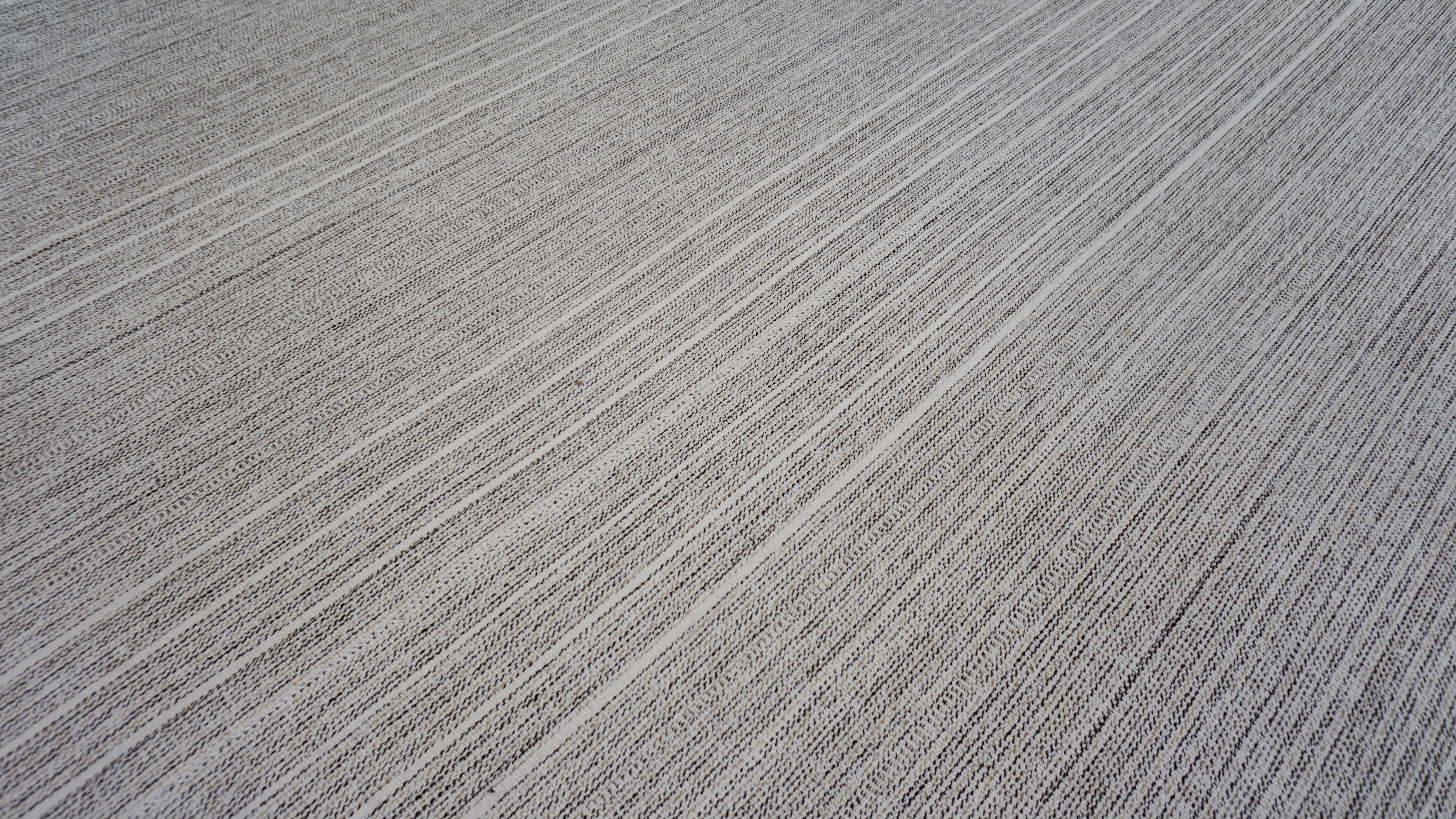 This handwoven wool kilim showcases the purity of minimalist design rooted in centuries-old weaving traditions. The palette is a sophisticated interplay of soft gray, ivory, beige, and charcoal woven into vertical pinstripes that quietly echo the