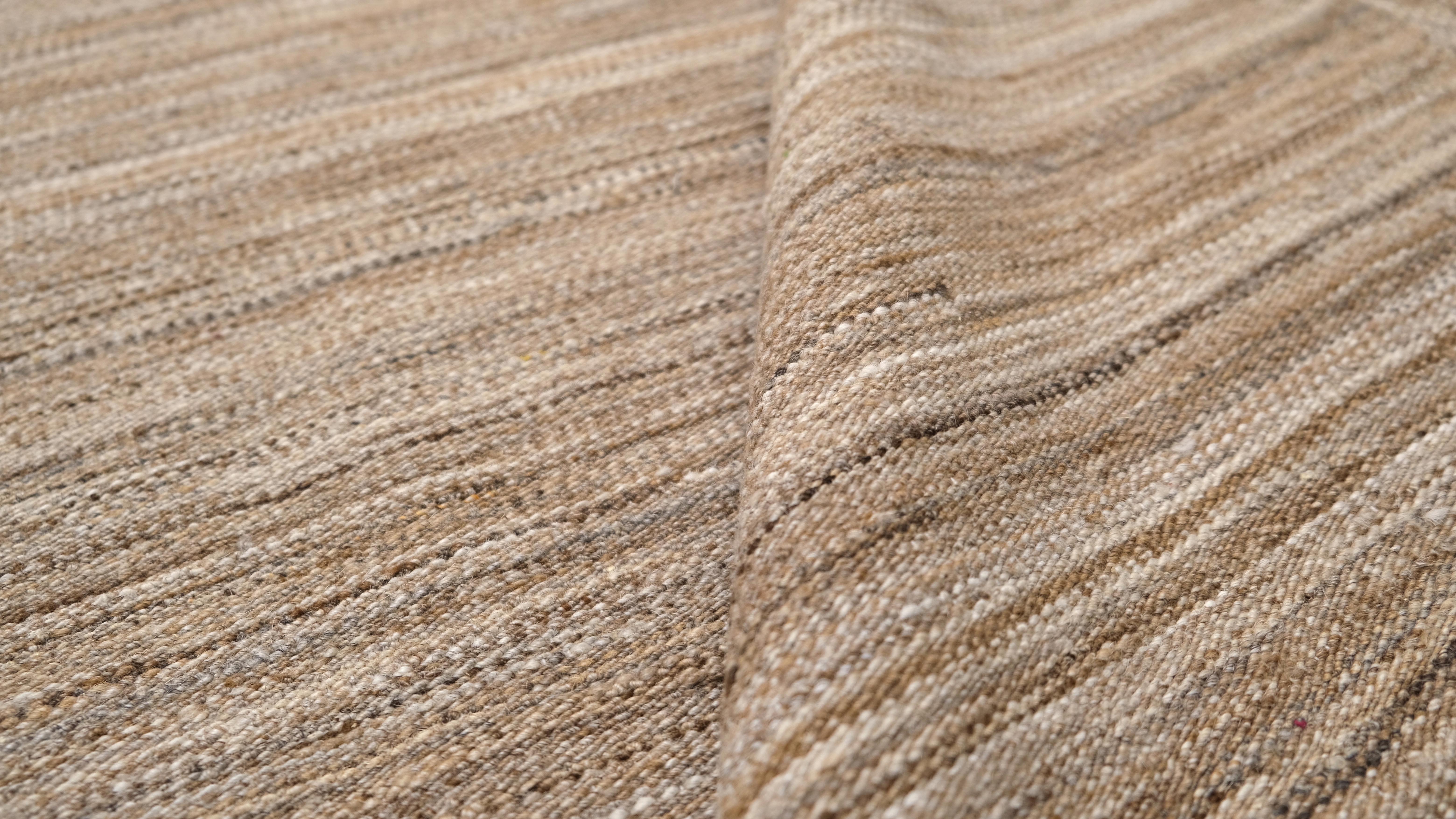 Dieser handgewebte Wollteppich ist ein Paradebeispiel für zeitgenössischen Minimalismus in Verbindung mit traditioneller Handwerkskunst. Mit seinem dezenten Streifenmuster in natürlichen Beige- und Sandtönen strahlt dieser Teppich eine schlichte