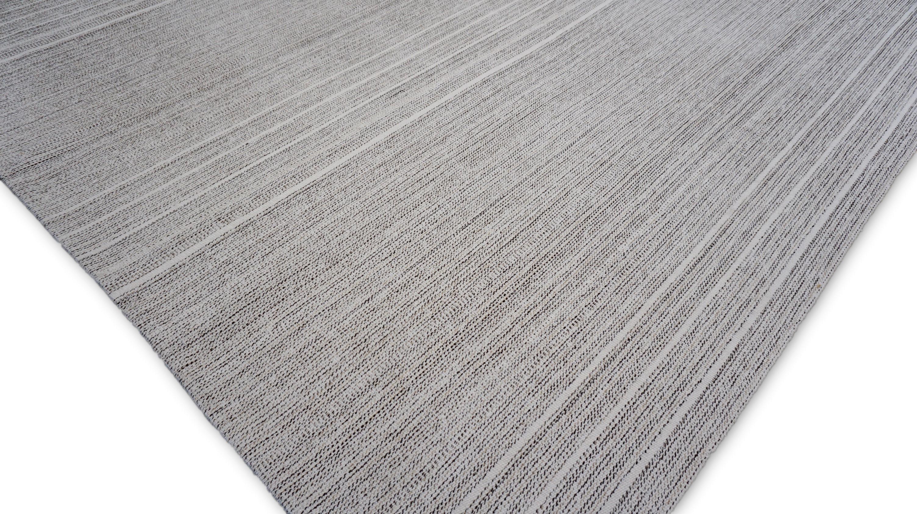 Dieser handgewebte Wollteppich zeichnet sich durch ein schlichtes und minimalistisches Streifenmuster in verschiedenen Grautönen, Weiß und Elfenbein aus. Das subtile, lineare Design schafft ein Gefühl von Tiefe und Bewegung und macht es zu einer
