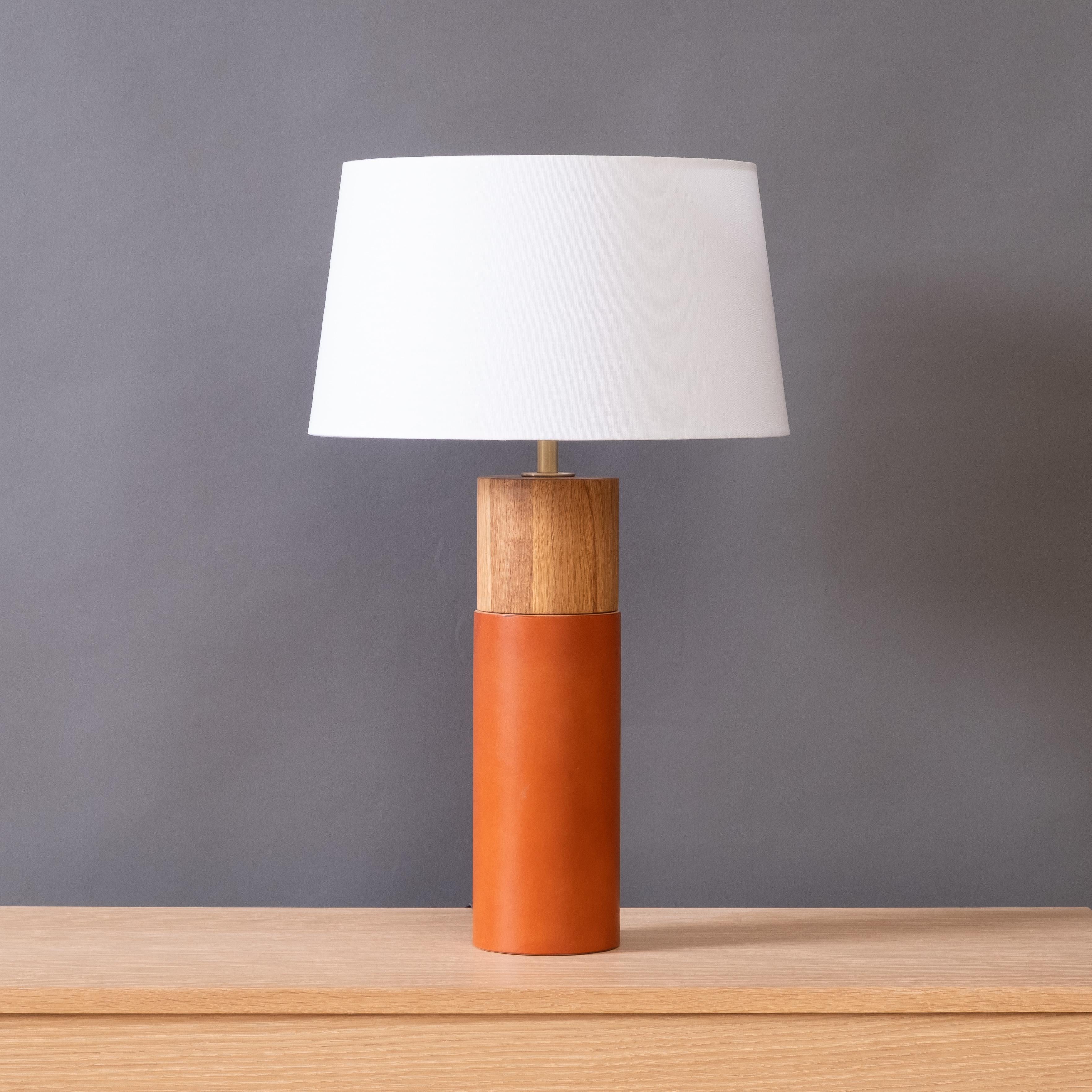 The Capsule table lamp combines minimalist forms with richly textured natural materials. Thick English Bridle leather wraps the lower portion of the turned wood body, contrasting with the oiled wood exposed in the top portion. The satin-finish brass