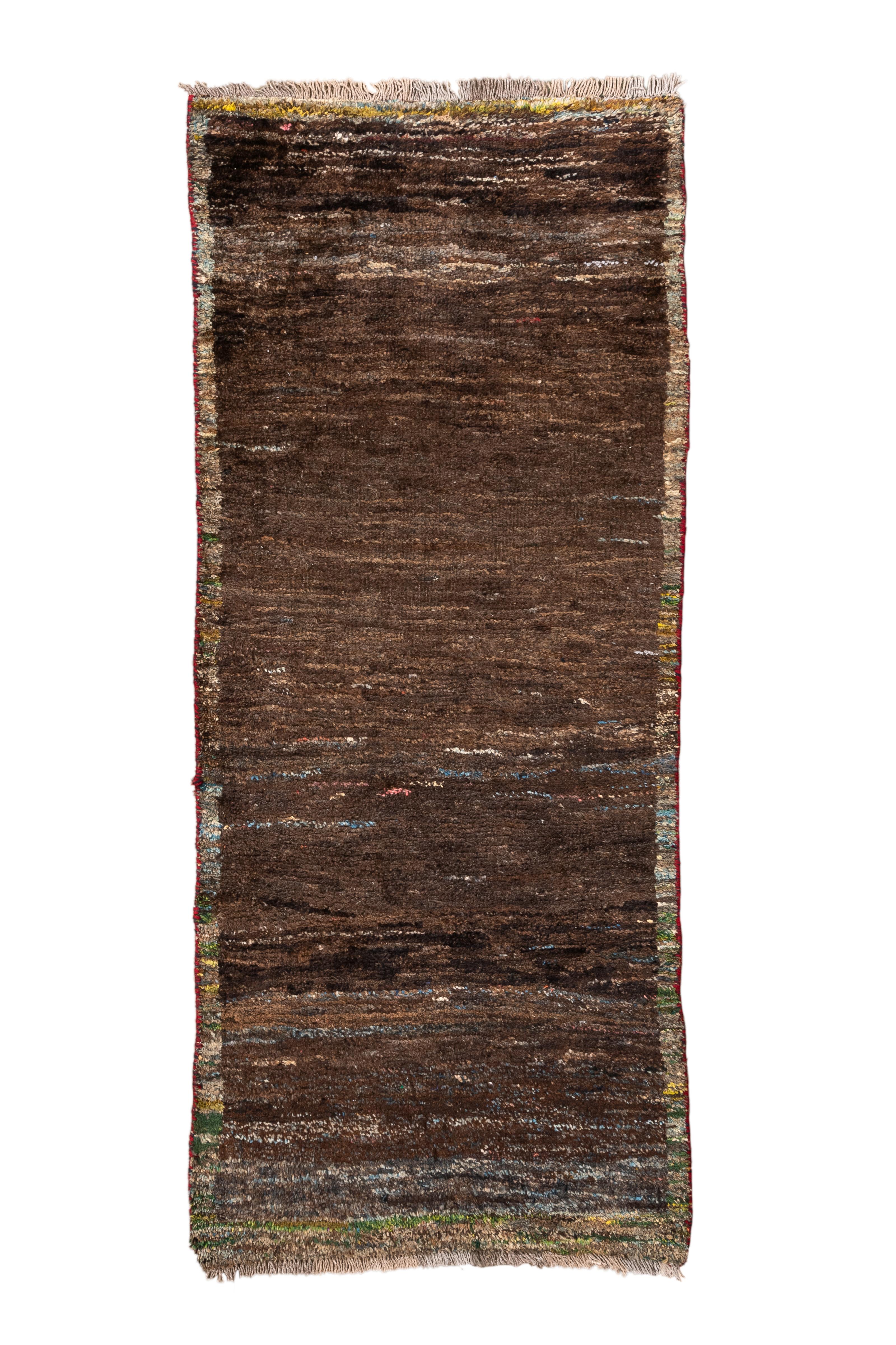  A deeply textural and meditative Gabbeh rug in rich shades of naturally dyed brown, accented subtly with multicolor speckling and irregular variegation. The minimalist field is framed by faint, painterly bands at the edges and ends, evoking the raw
