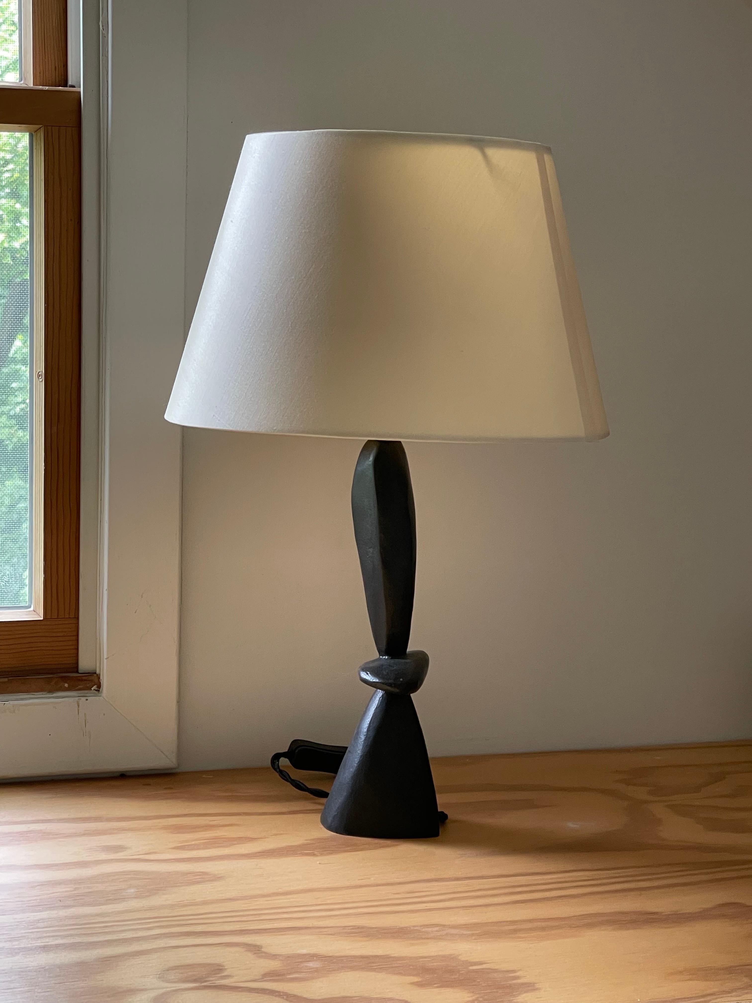 Menine is a sculptural table lamp by Jacques Jarrige, cast in bronze and composed of three interlocking elements. A rounded, pebble-like form rests horizontally, cradled between two vertical supports that rise with quiet presence. The composition is