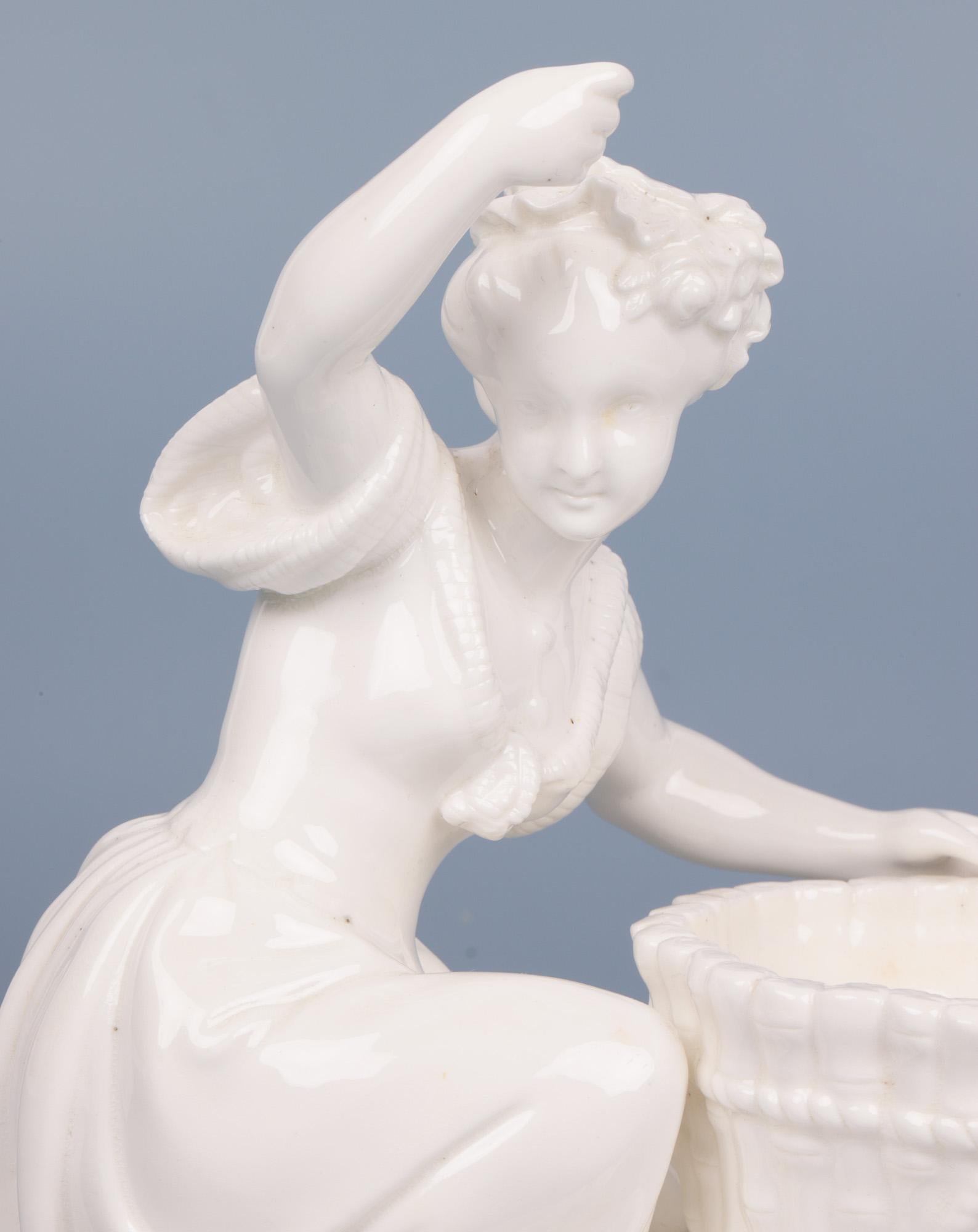 Fino centro de mesa antiguo de porcelana blanc de chine de Minton con una joven doncella con una cesta que data de alrededor de 1862. El centro de mesa se alza sobre una base ovalada y está formado por una joven arrodillada junto a una cesta. Está