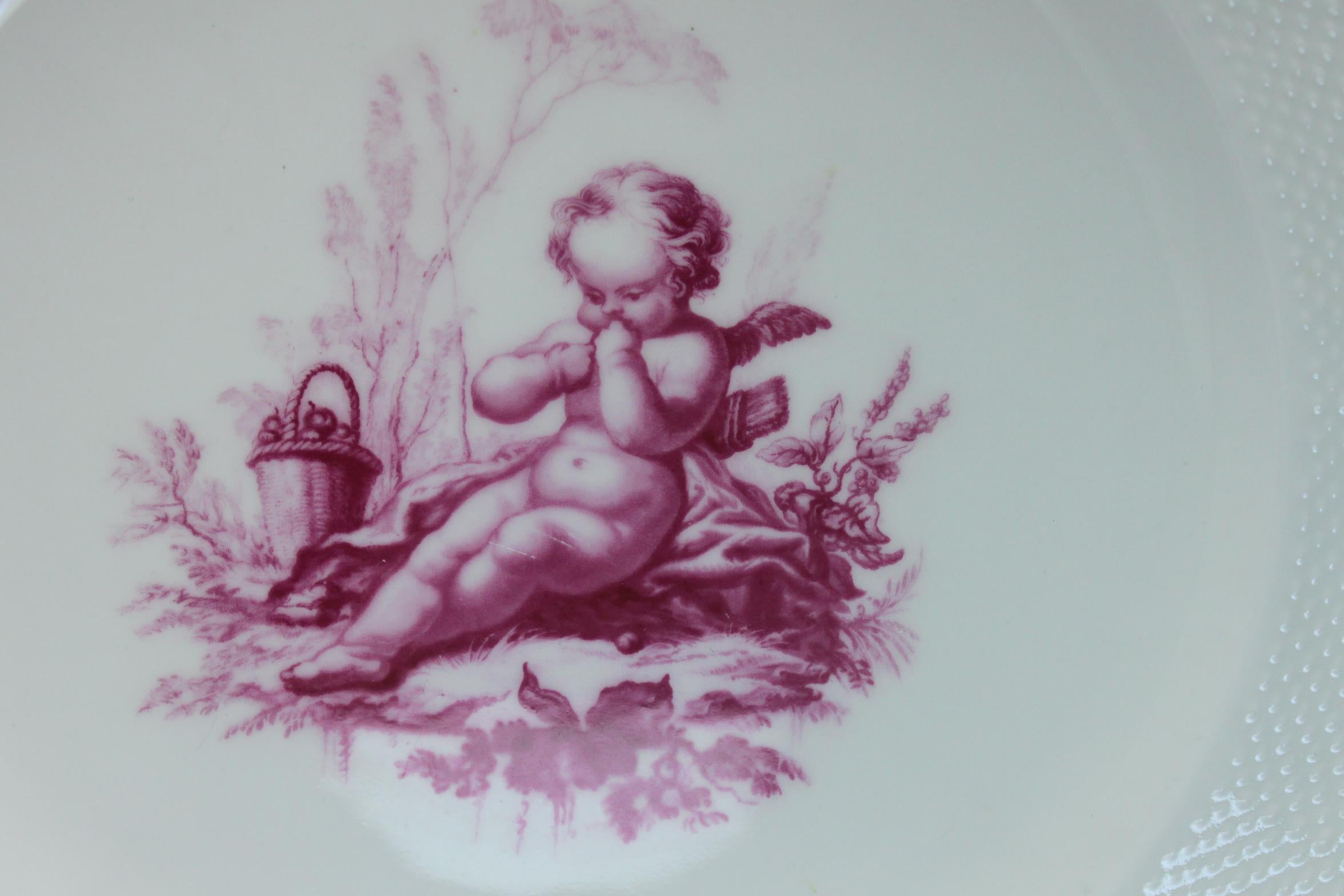 This Minton plate is decorated very simply with a central hand coloured transfer print of a cherub in puce (pattern G1533) The outer rim features a very fine basket weave moulding and the edge is finished with gilding. It was made in 1874 for the