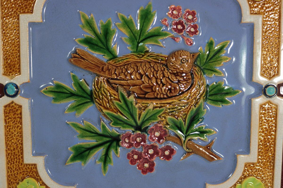 Minton Majolica tile which features a nesting bird, amongst foliage and flowers. Colouration: indigo, green, brown, are predominant. The piece bears maker's marks for the Minton pottery.